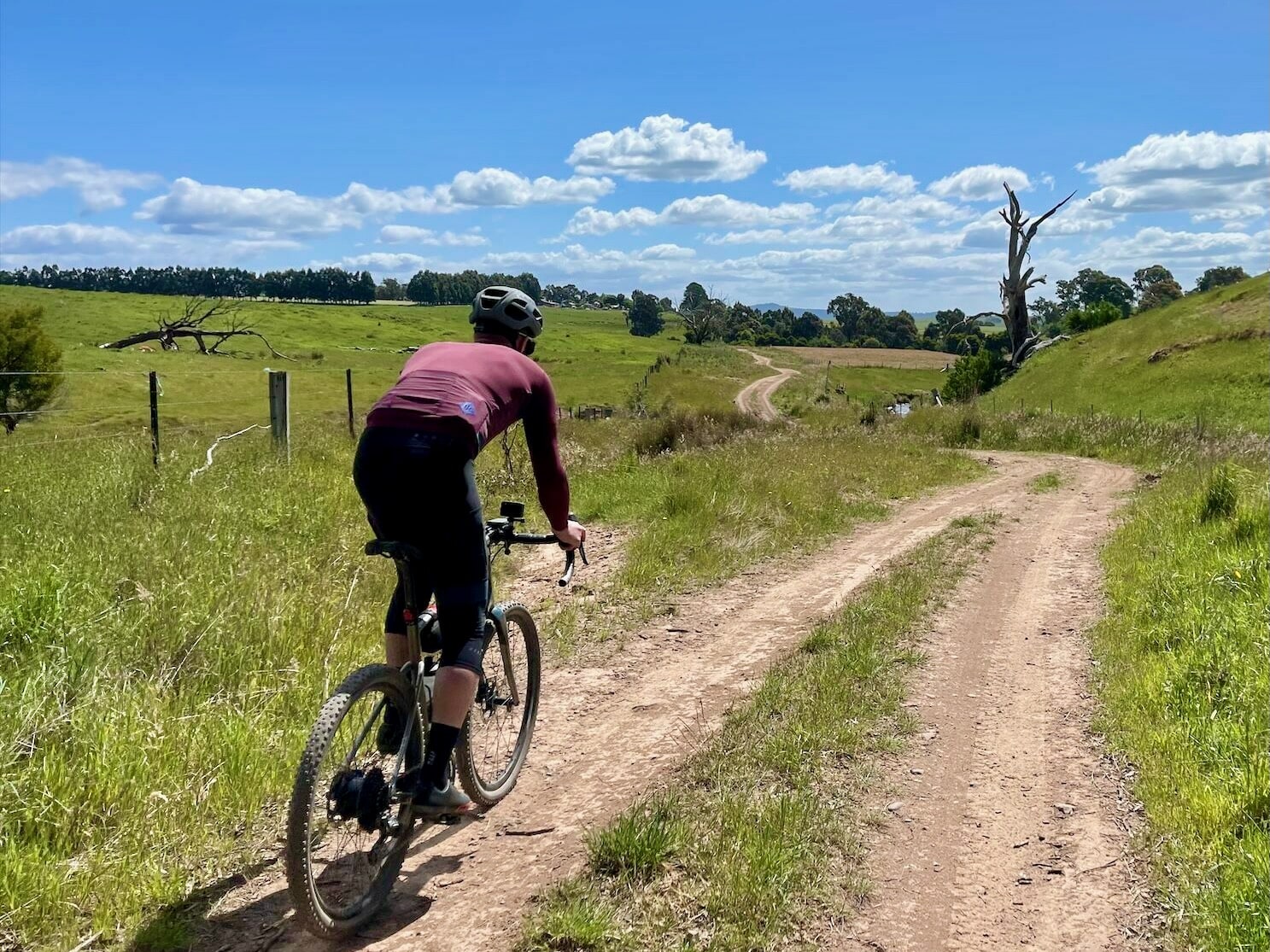 Cyclist riding on a dirt track which runs through green lush open farmland  on a sunny day
