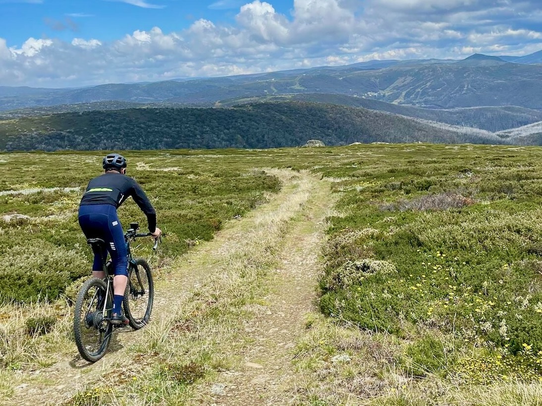 Cyclist riding a gravel double track through the high alpine with Falls Creek resort in the distance 