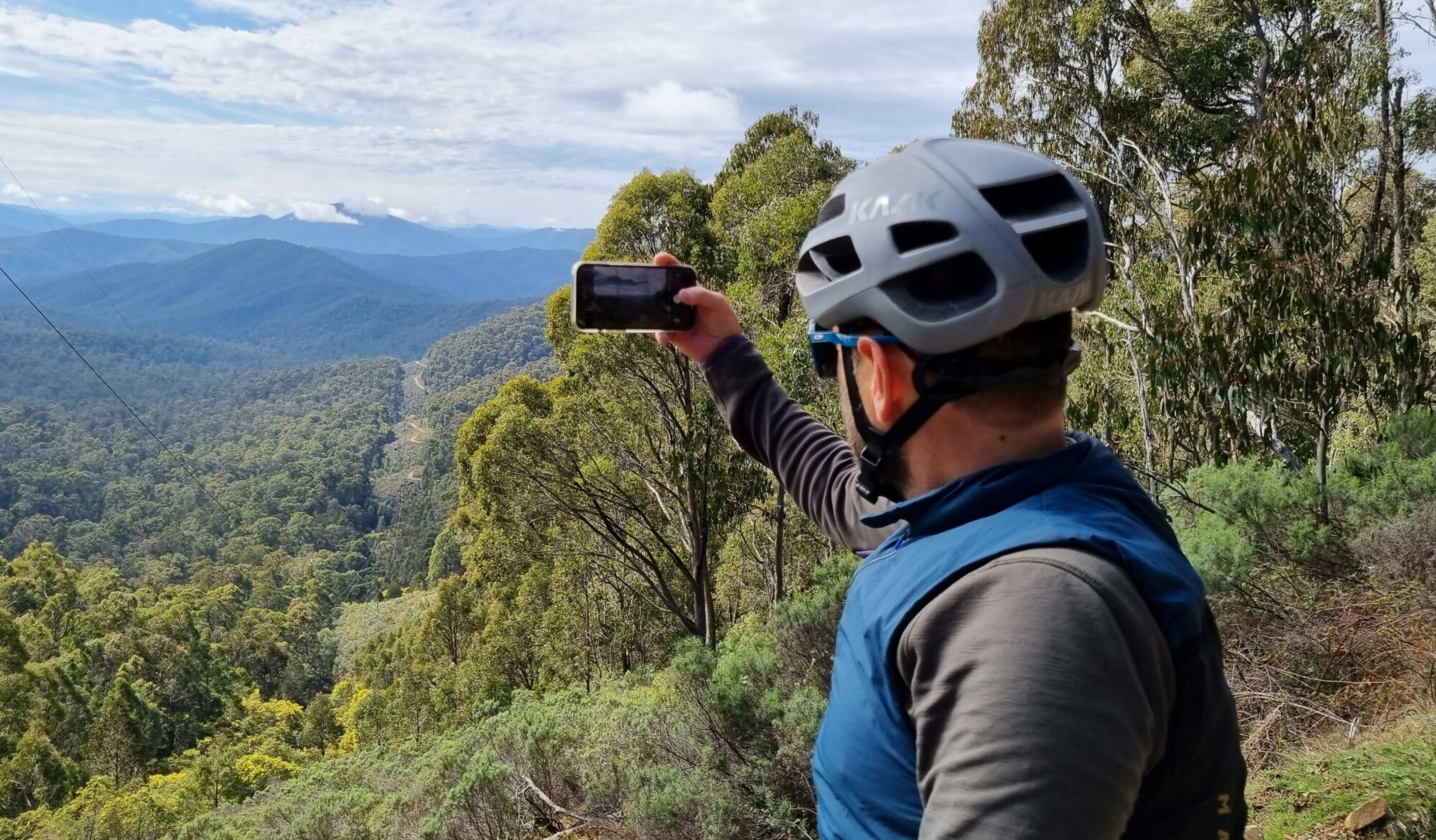Cyclist stopped on the side of the gravel road taking a photo of the surrounding mountains and valleys which are covered with native bushland 