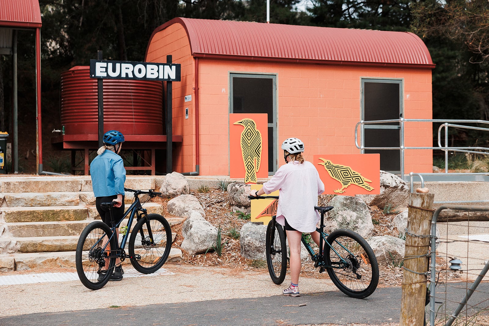 Two female cyclists in front of a building signed 'Eurobin' with stone steps and indigenous artworks of native animals.