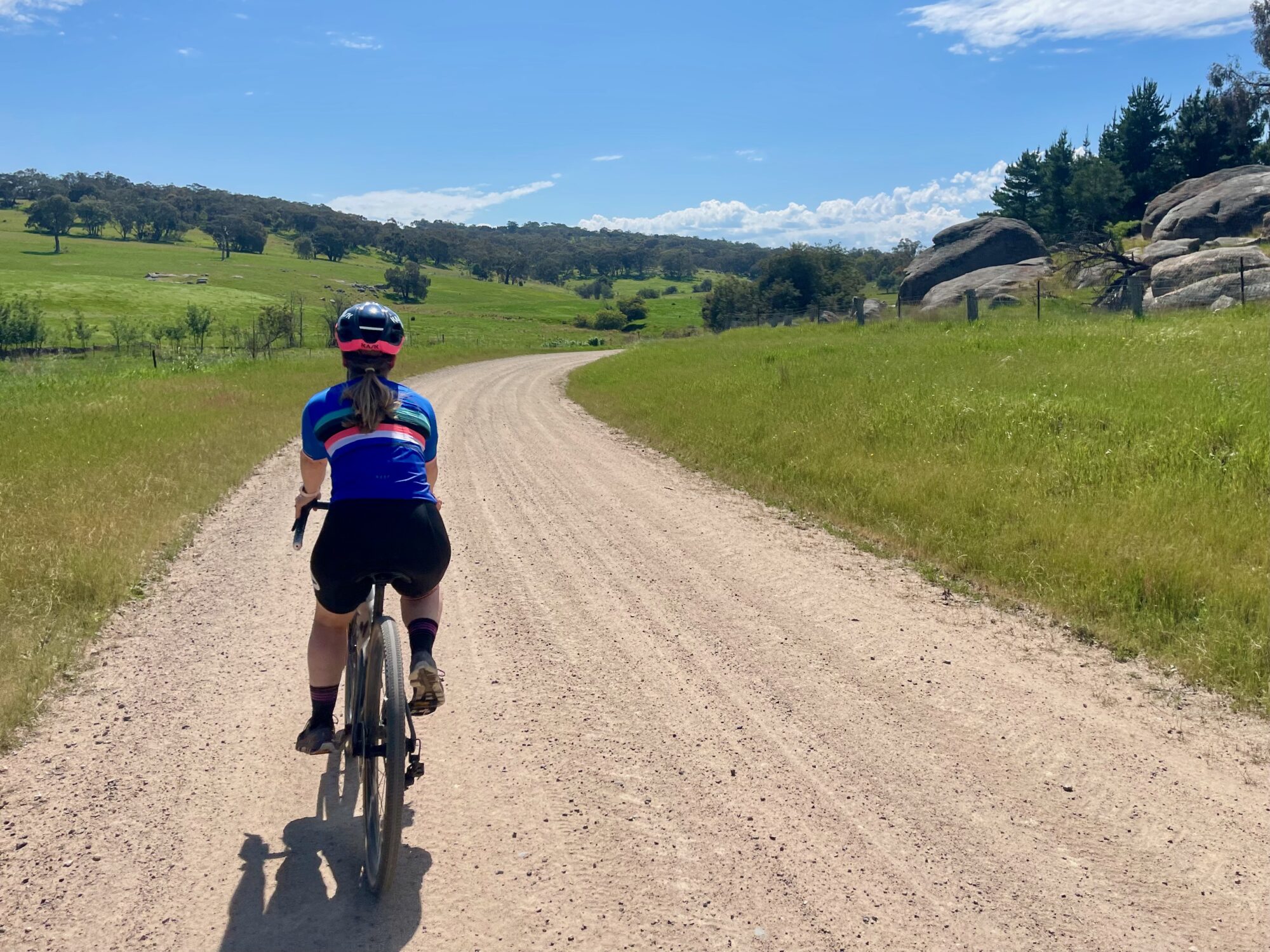 A female cyclist riding under blue skies on a smooth gravel road that is winding through open farmland and large boulders on the side of the road with native bushland in the background 