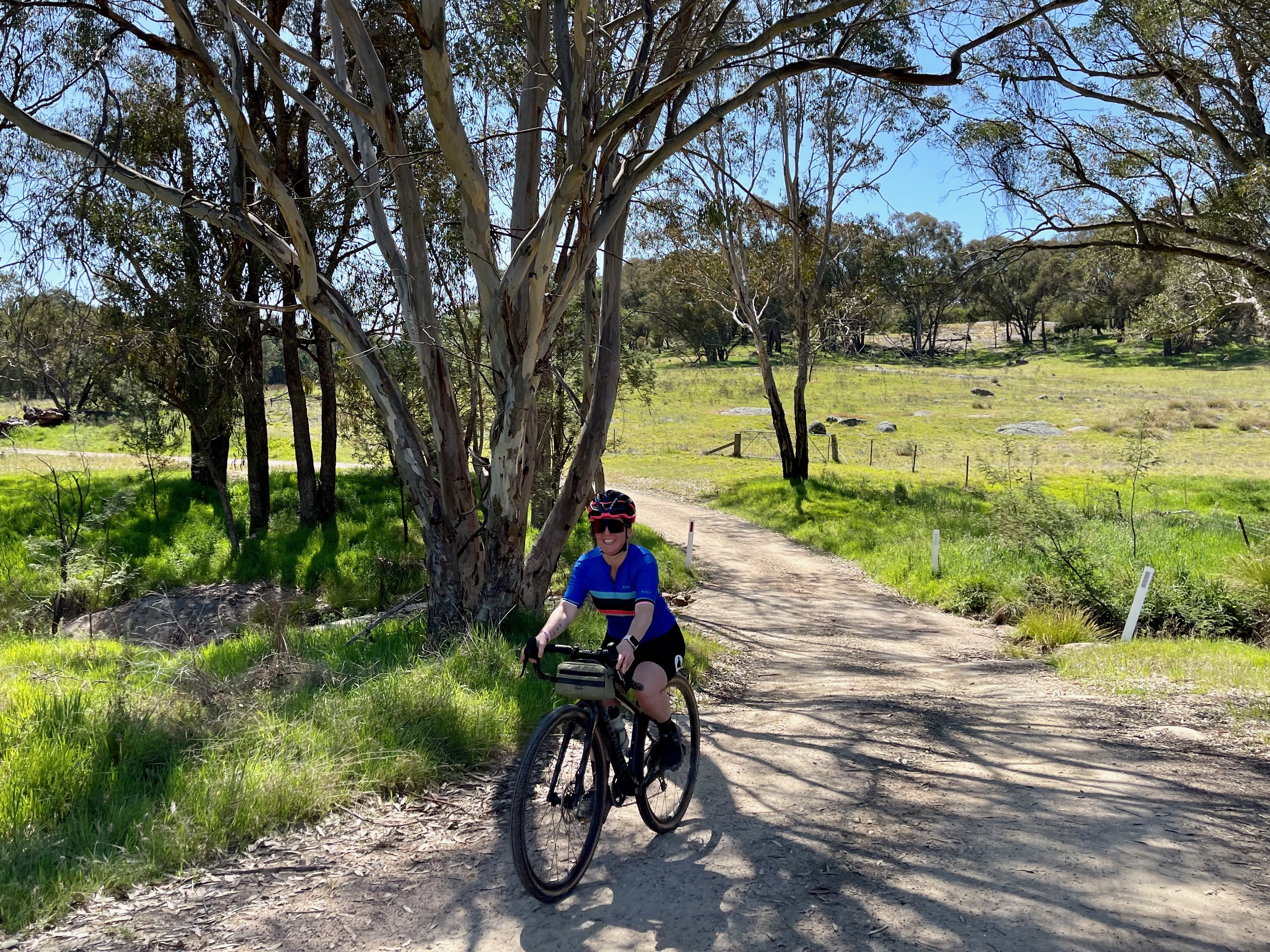 A female cyclist riding under blue skies on a smooth gravel road that is winding through open farmland and native bushland