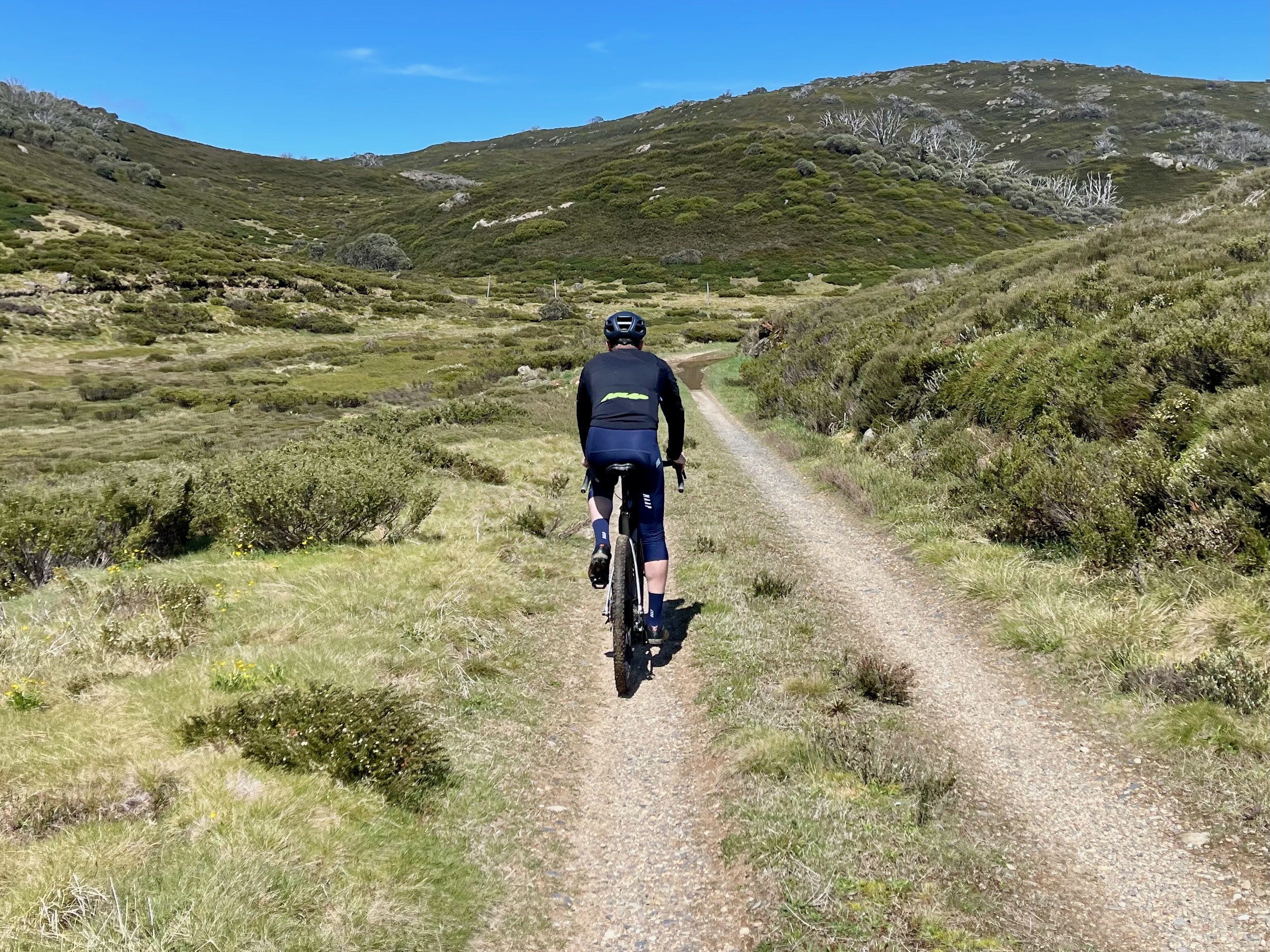 Cyclist riding on a smooth gravel double track at the base of surrounding hills and surrounded by lush green tussock grass on a sunny day