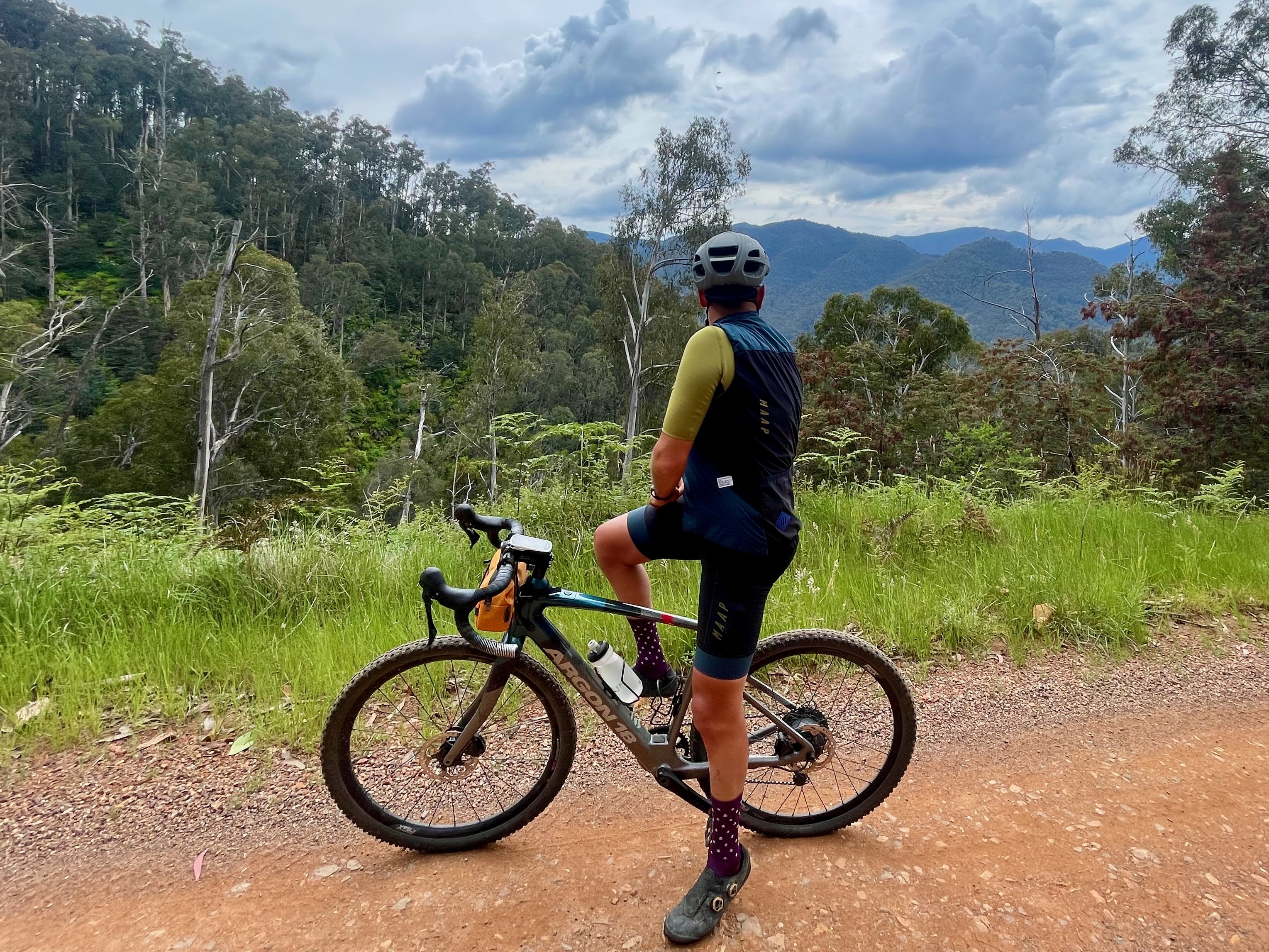 A cyclist stopped on a smooth dirt road looking at the view of surrounding native bushland covered mountains and valleys