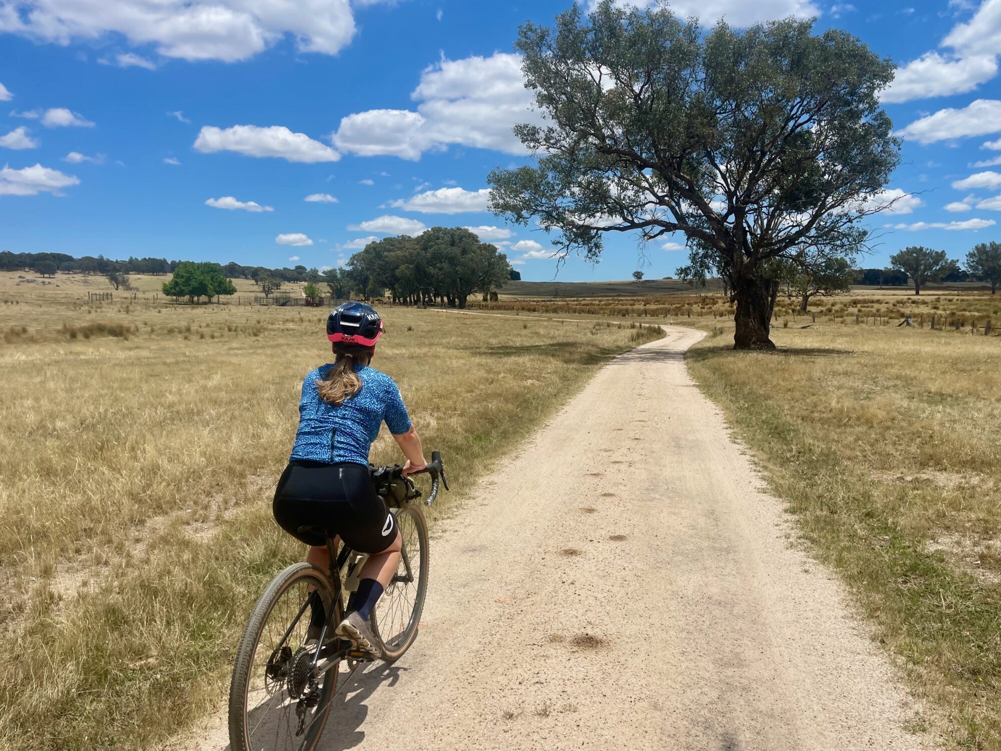 A female cyclist riding on a smooth champagne crushed granite gravel road through open farmland with scattered native trees lining the gravel road under blue skies and fluffy clouds