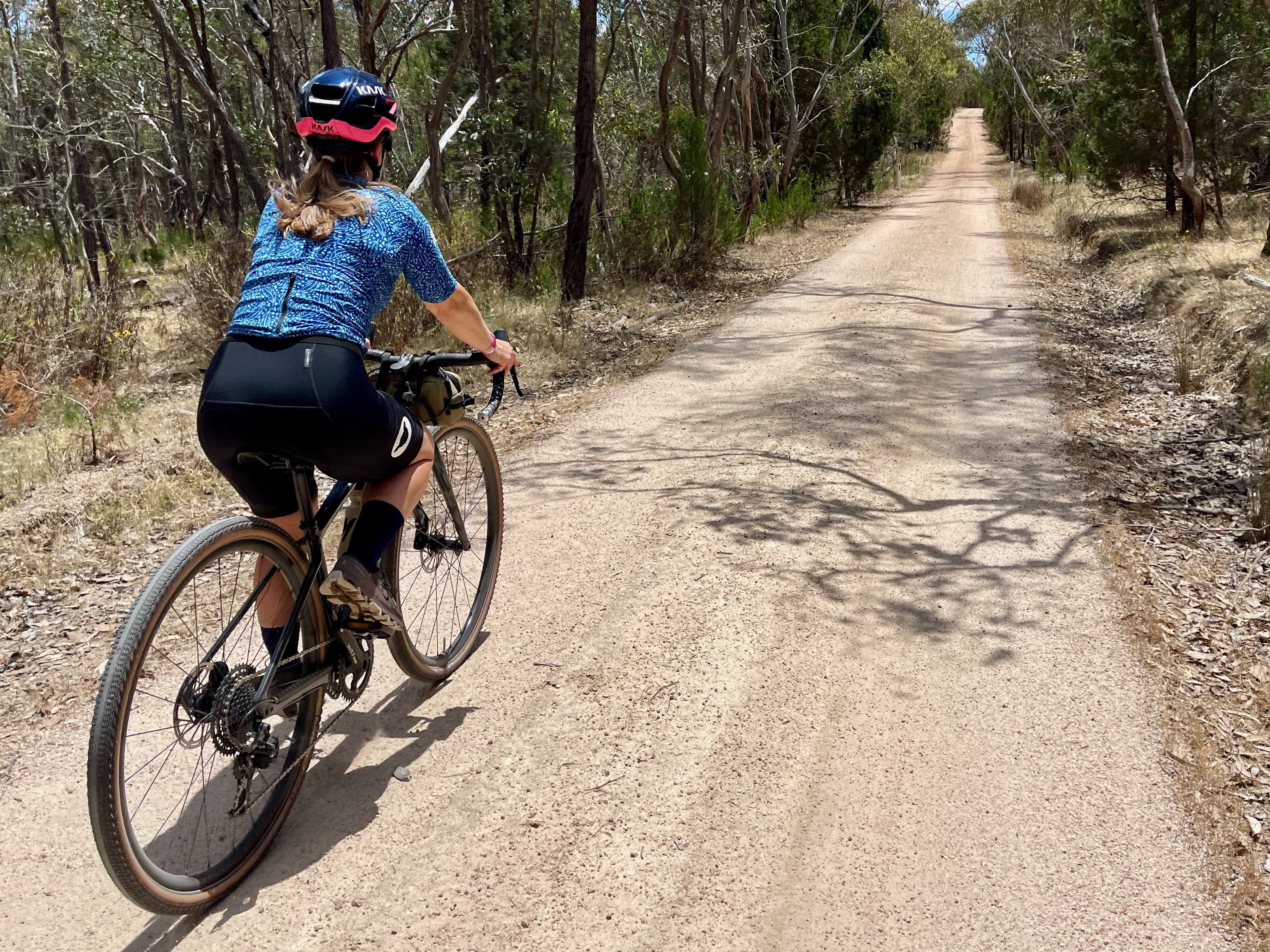 A female cyclist riding on a smooth champagne crushed granite gravel road through open farmland with scattered native trees lining the gravel road surrounded by native bushland on a sunny day