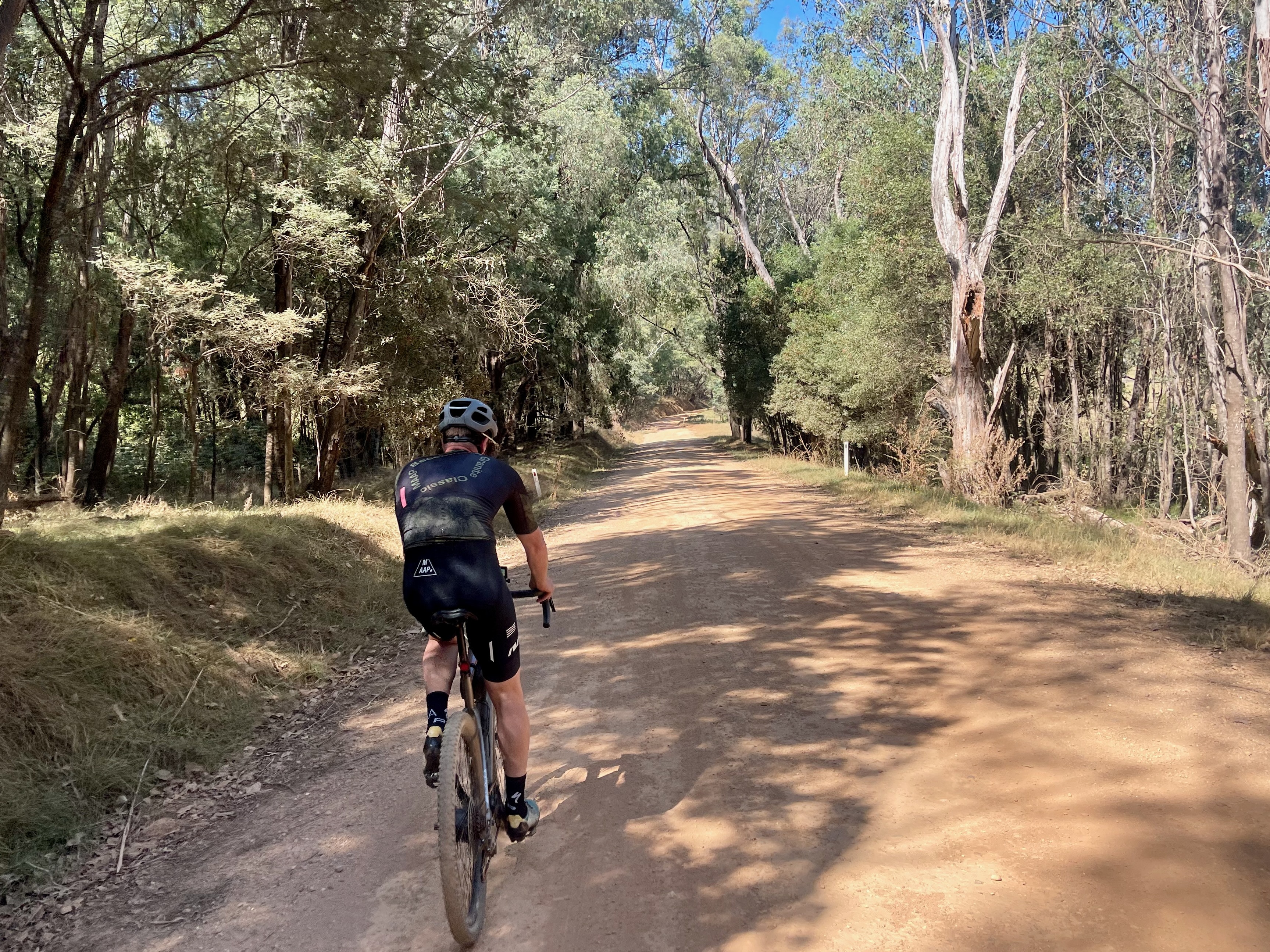 Cyclist riding on a quiet gravel road through native bushland with farmland and mountains in the background on a sunny day
