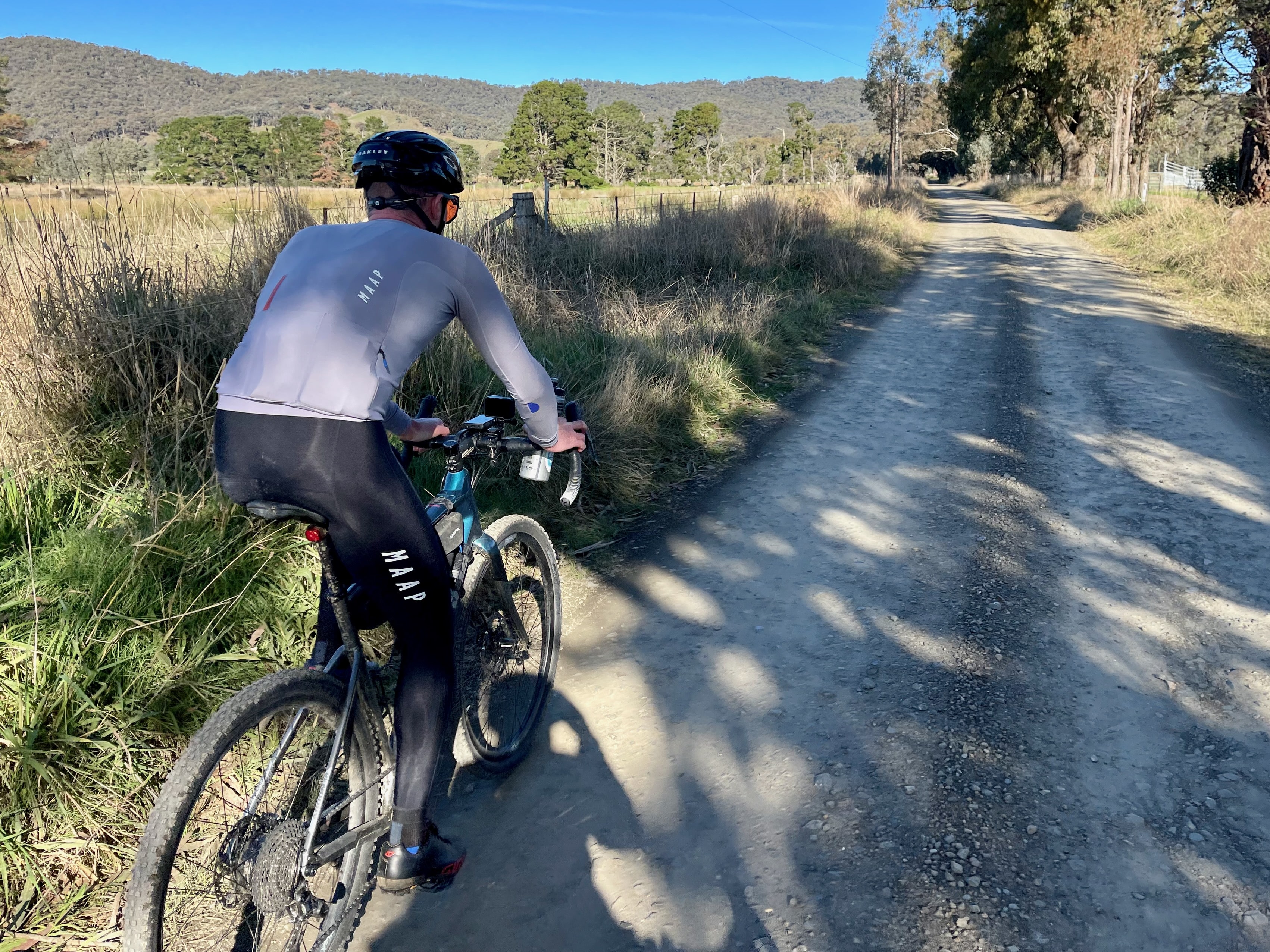 A cyclist riding on a gravel road with open farmland and native bushland in the background on a sunny day