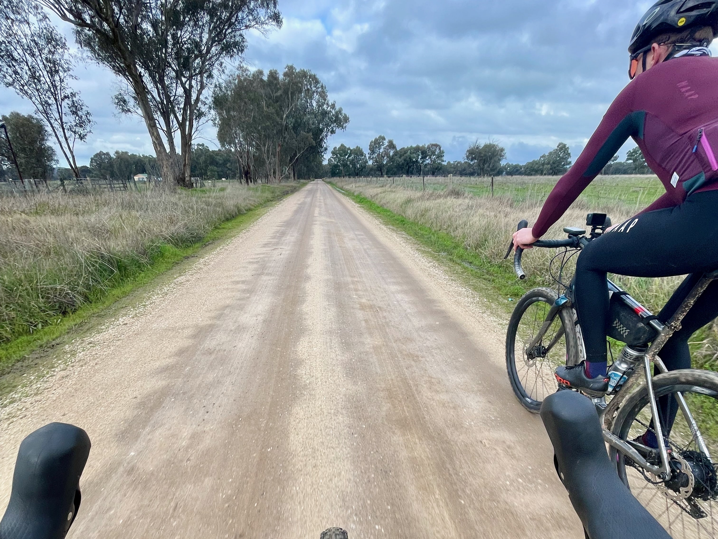 Two cyclists gravel riding on a smooth dirt road surrounded by open farmland and native bushland in the background