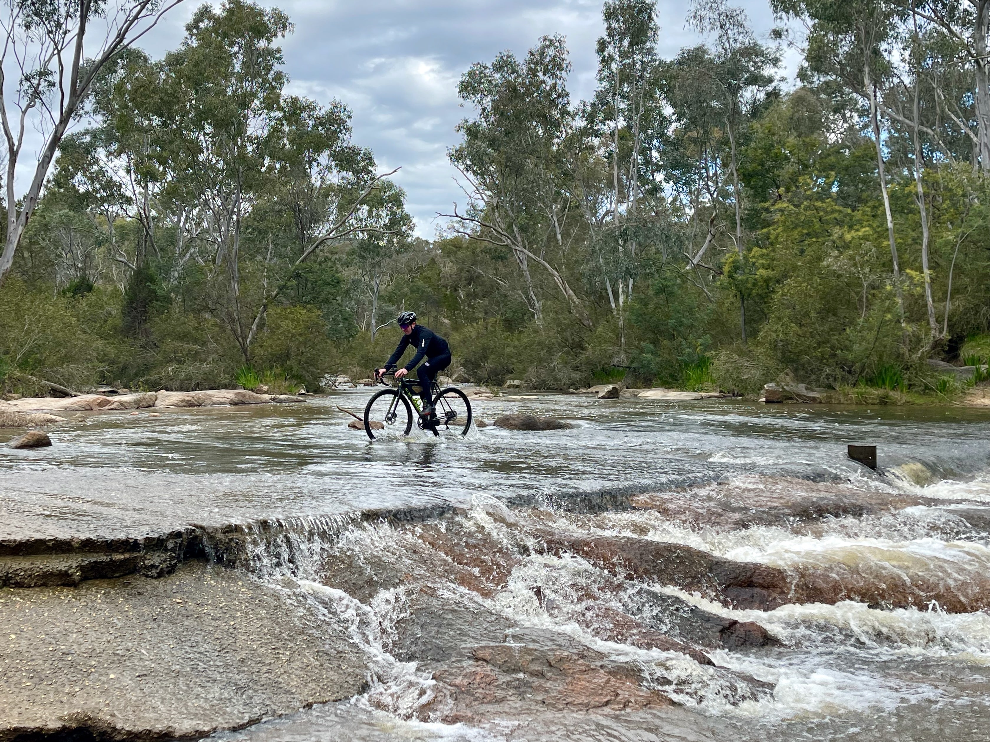 A cyclist rides across a shallow river crossing with native bushland surrounding