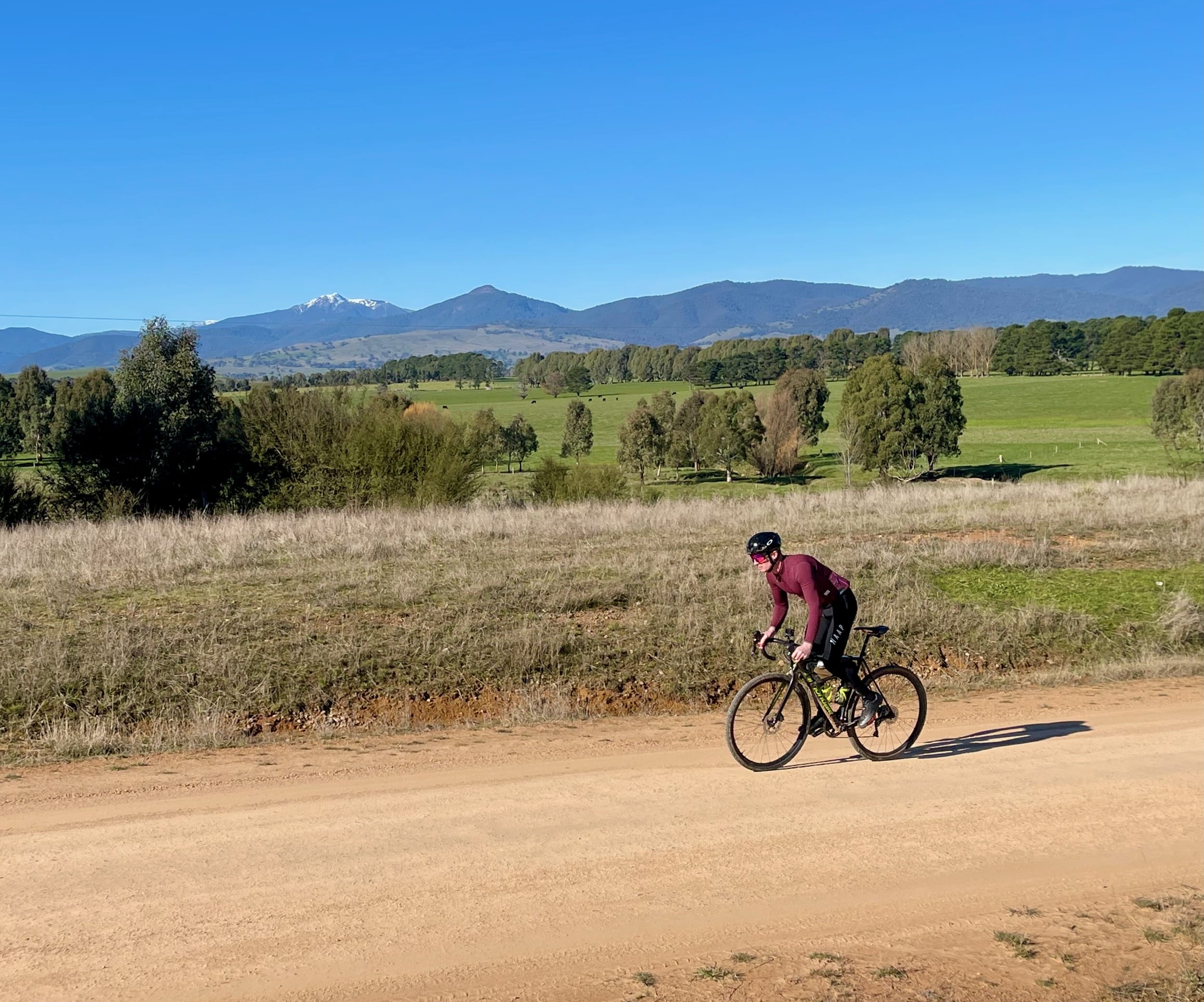 Cyclist riding on a smooth gravel road through open farmland and snow capped moutains in the background on a sunny day