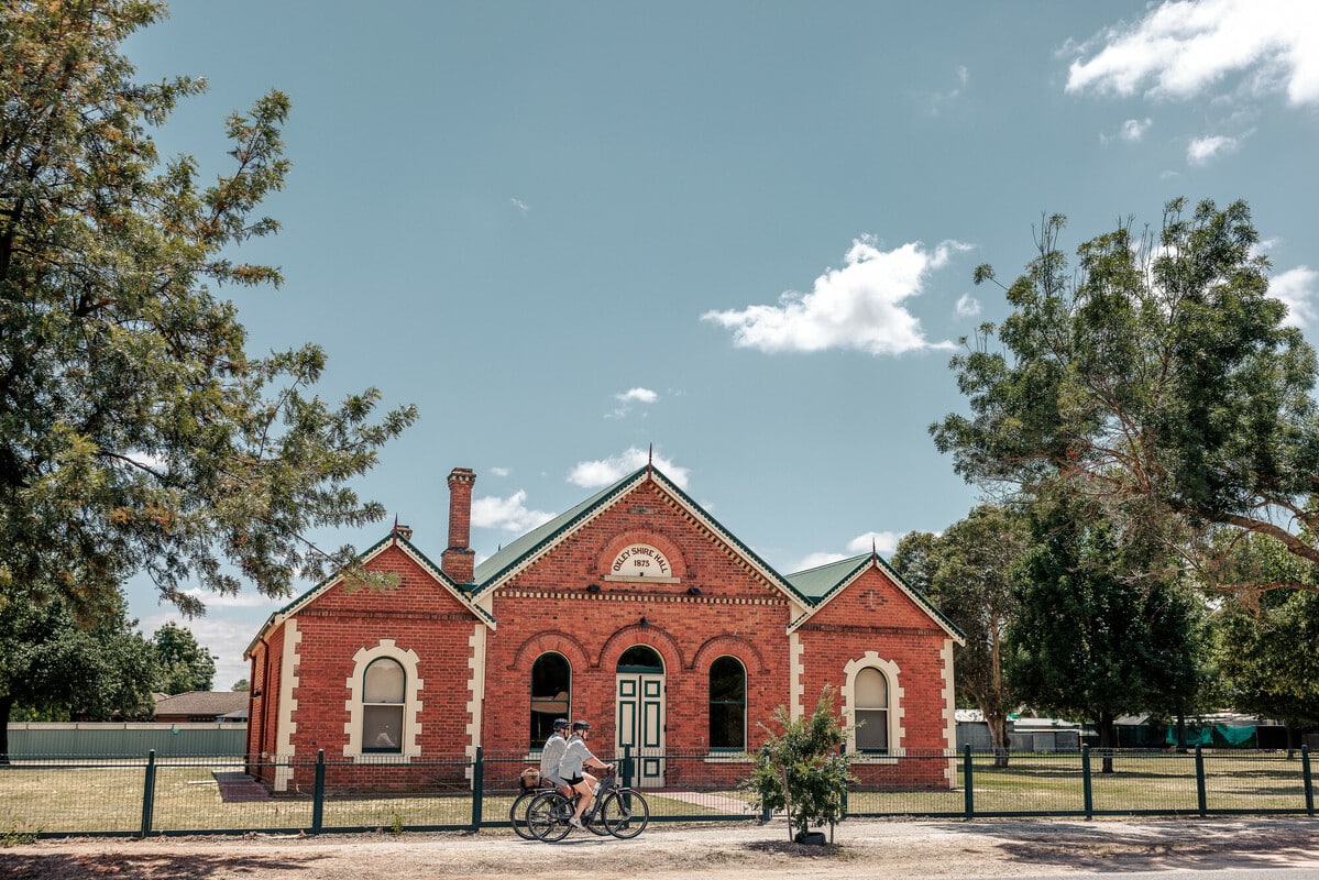 Two cyclists riding along a trail past an 1870's brick hall. 