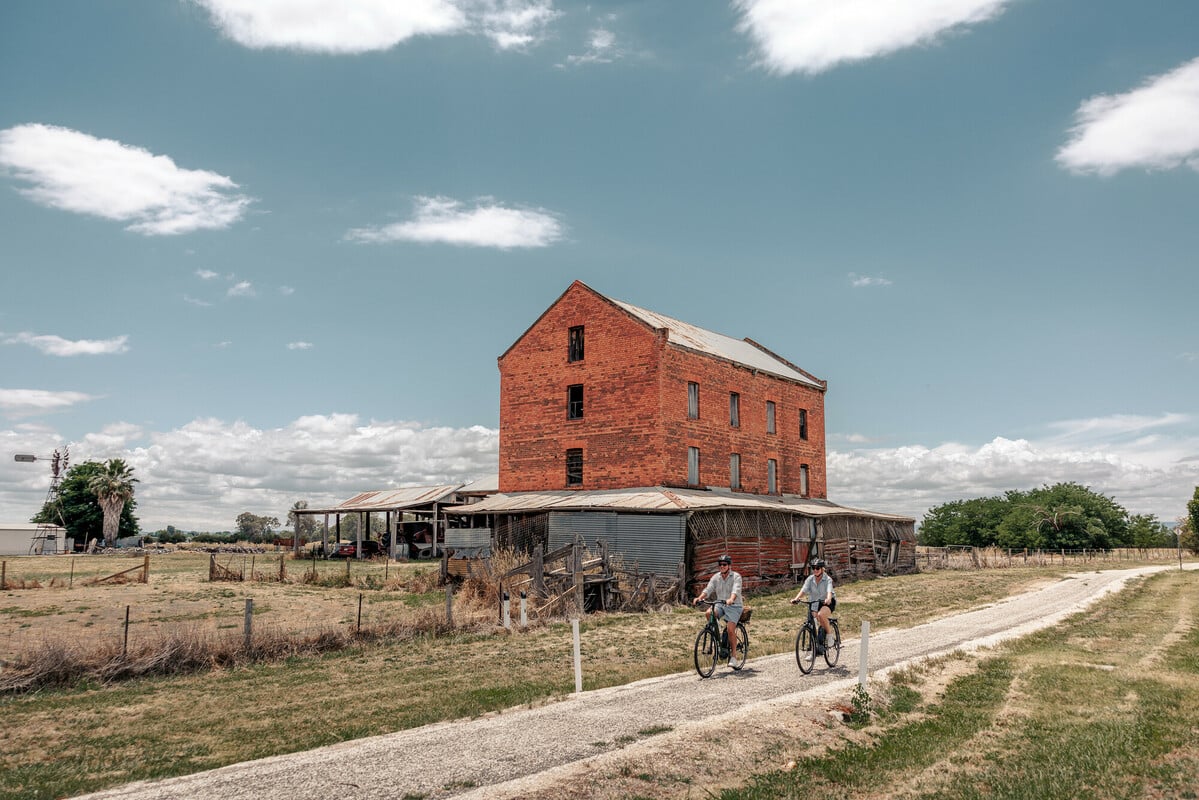 Two cyclists riding long a path past an historic brick building in a paddock.