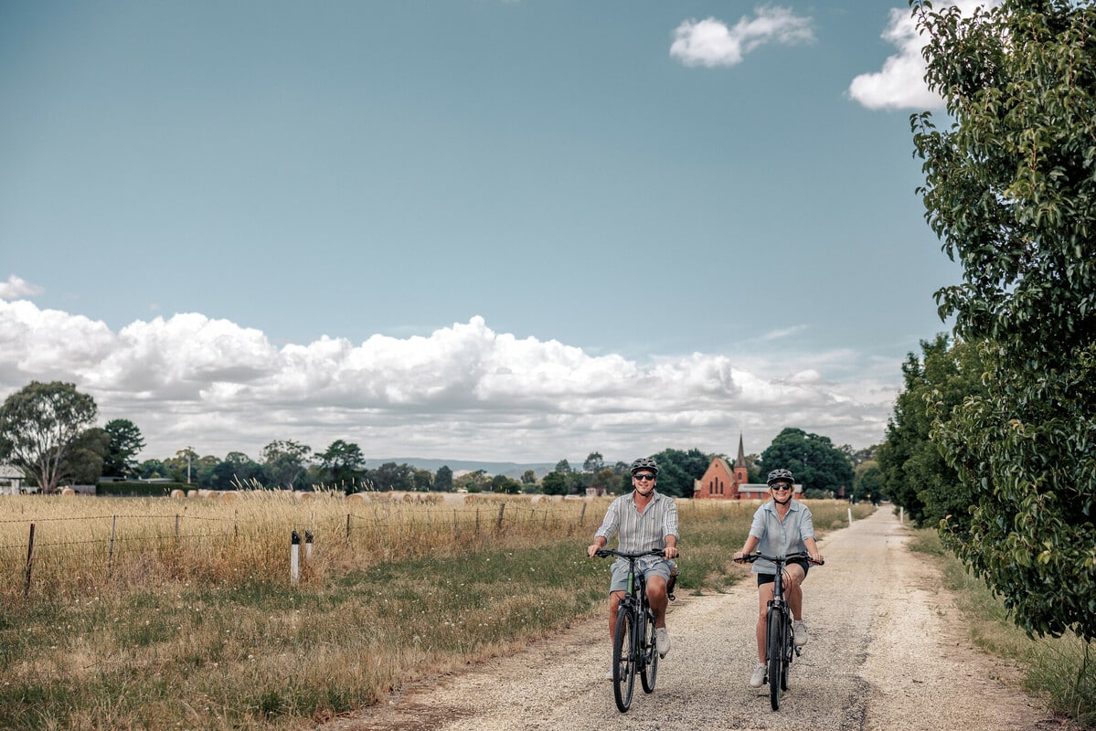 Two cyclists riding past a paddock and luscious trees with a brick church in the distant background.