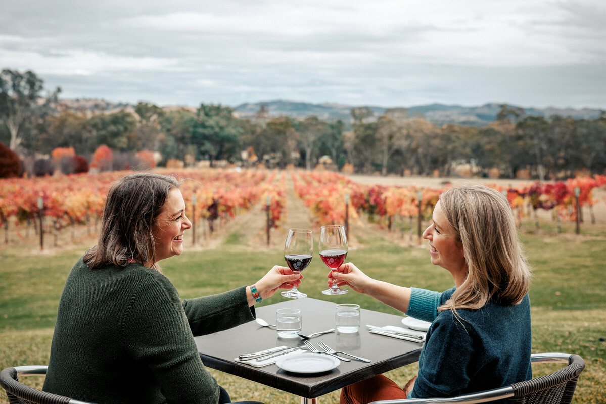 Two women cheers-ing prosecco and enjoying canapes at an alfresco dining table with an autumnal vineyard behind them.