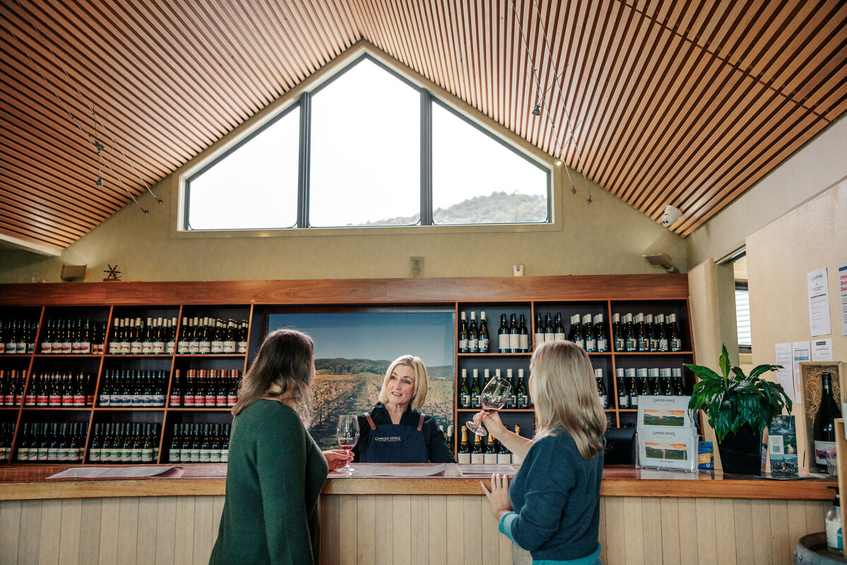 Two women enjoying a wine tasting with a female staff member in a cellar door with a pitched room and skylight, and a wall of wine bottles. 