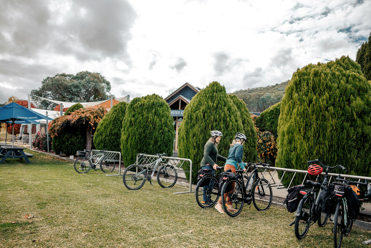 Two female cyclists popping their bikes into a long bike rack at a cellar door with leafy green trees.