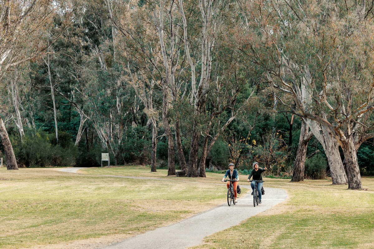 Two females riding bikes along a park trail surrounded by neat lawns and native Australian trees