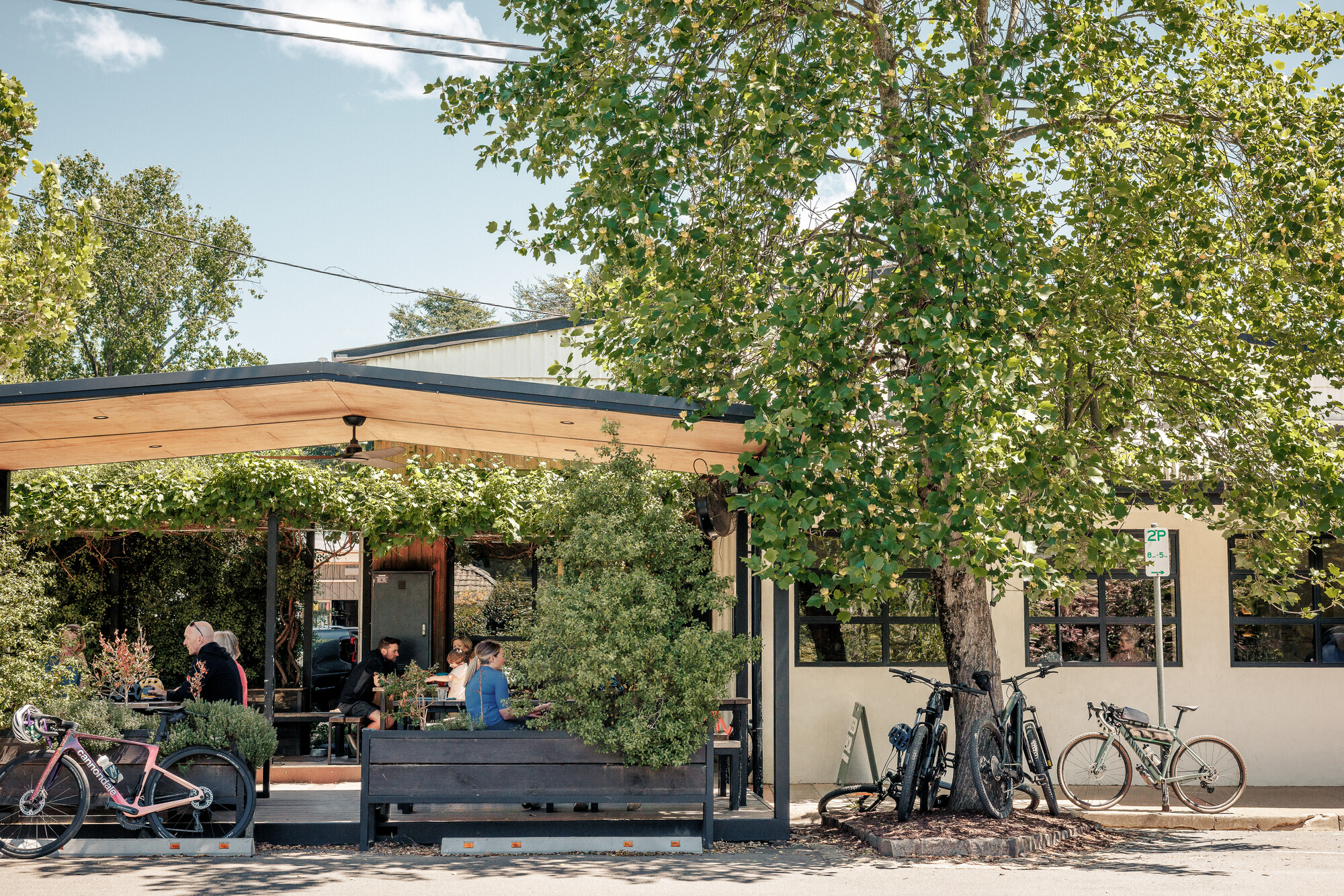 A multi-generational group of cyclists enjoying a leafy, alfresco courtyard attached to an industrial-chic looking coffee shop