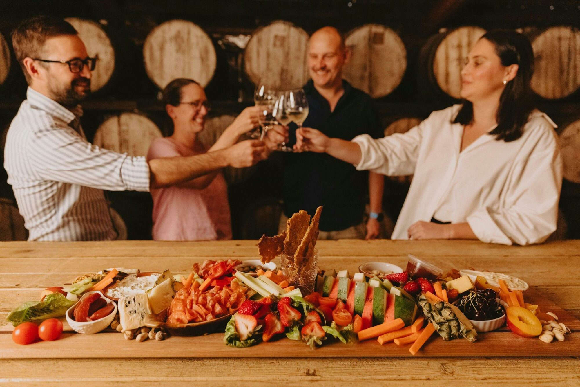 Four people cheering their wine glasses behind a large gourmet platter of local produce.