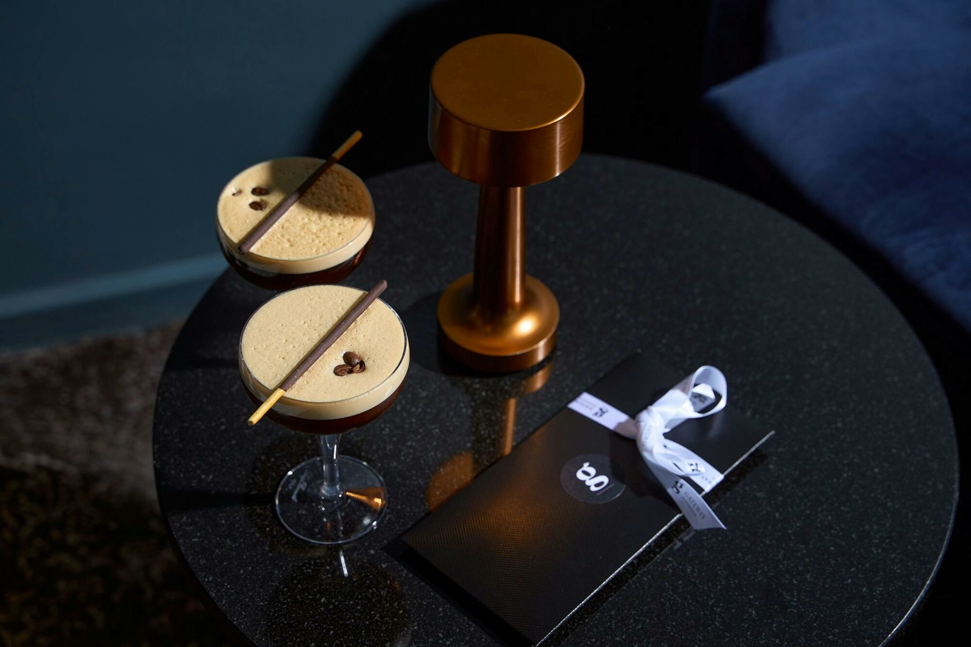 Two cocktails presented in candle lit bar with blue velvet sofas and gift with ribbon