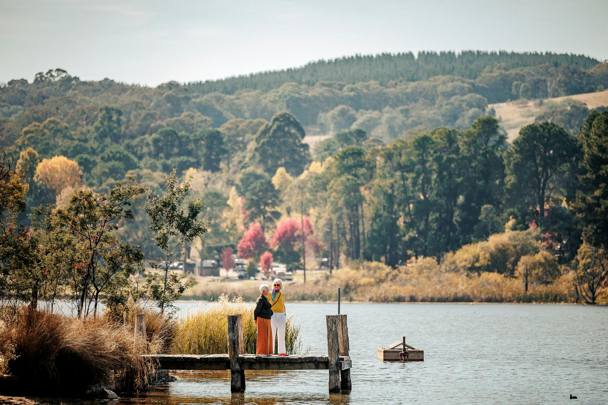 Two women on a jetty above a lake surrounded by hills and autumn trees