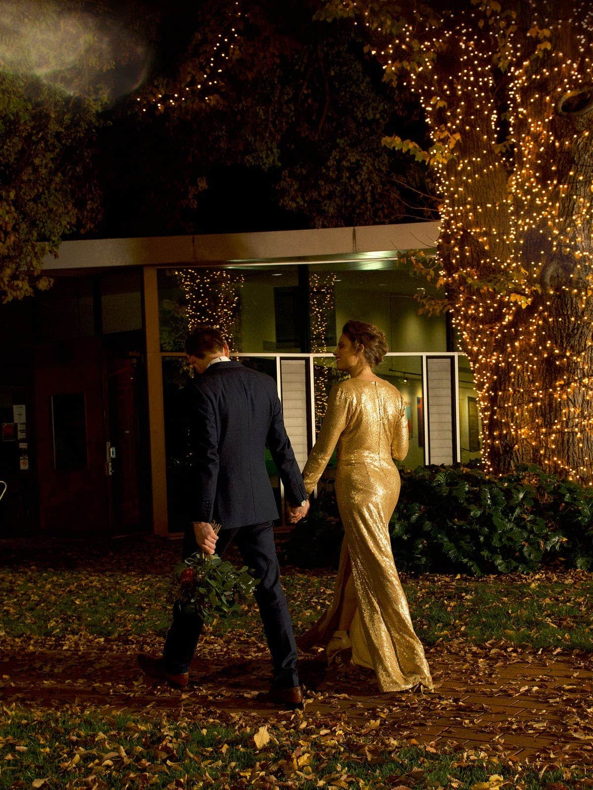 Couple dressed in after 5 attire walking towards entrance, fairy lights in tree,