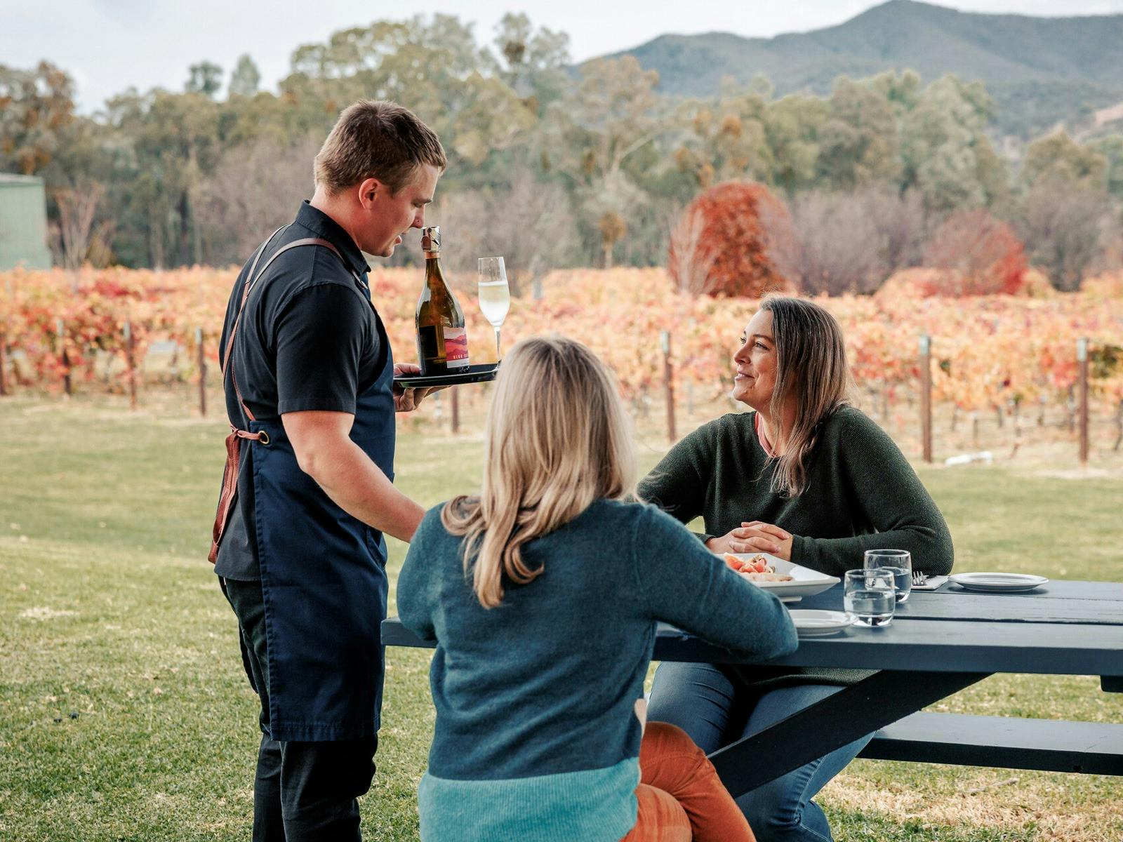 Man serving prosecco to two women sitting at a picnic table in front of autumnal vines