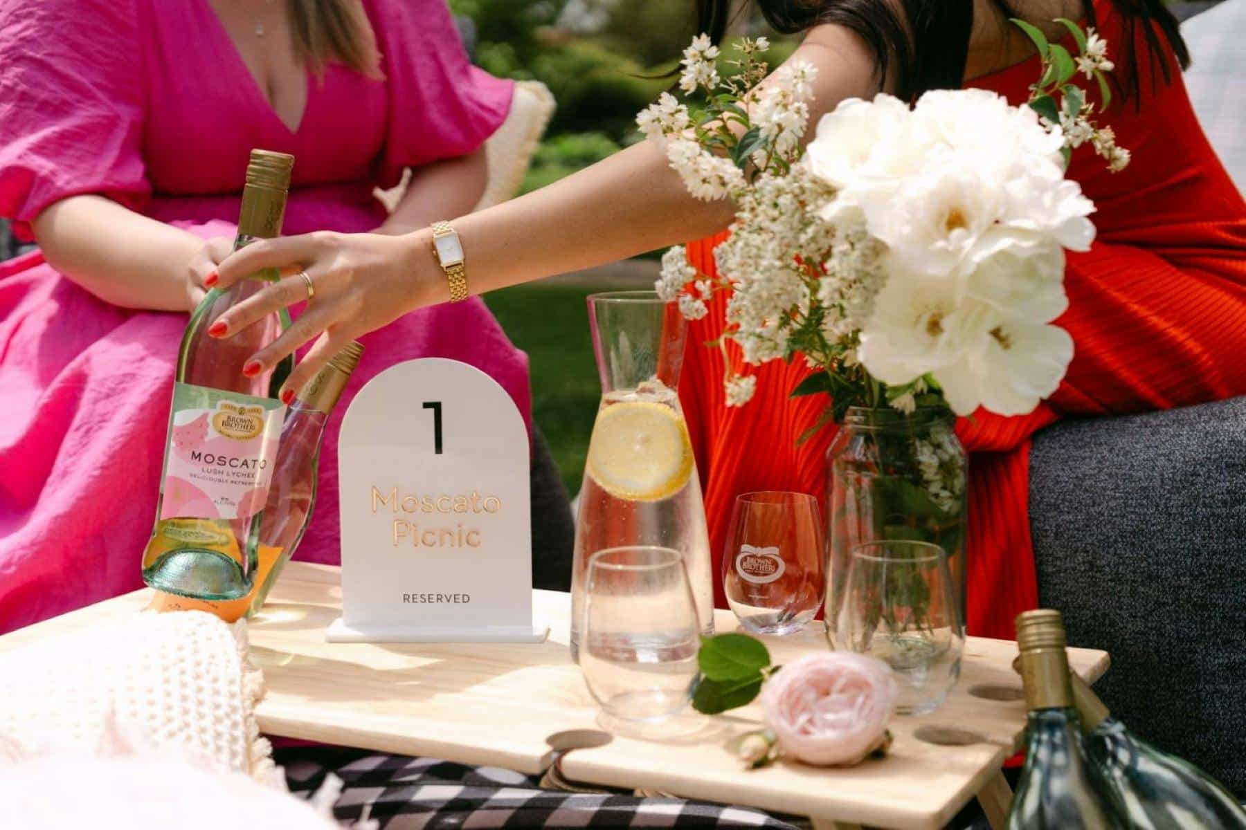 two people holding a wine bottle with glasses on a picnic table with a vase with white flowers
