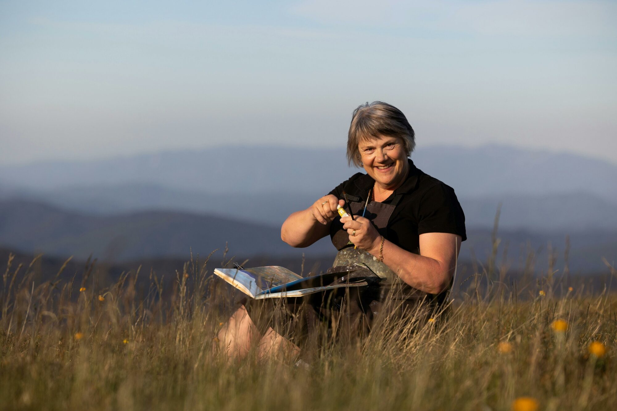 Artist Wendy Jagger sits among yellow wildflowers on a mountain top, with blue mountain ranges.