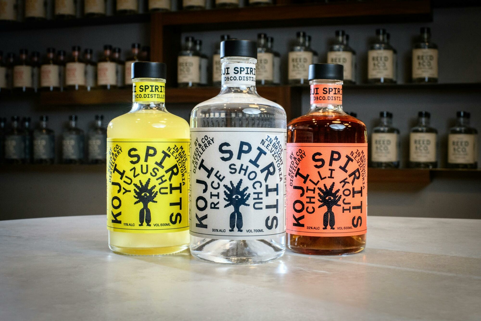 Three bottles of spirits with colourful labels sit on a bar with a shelf full of bottles behind.