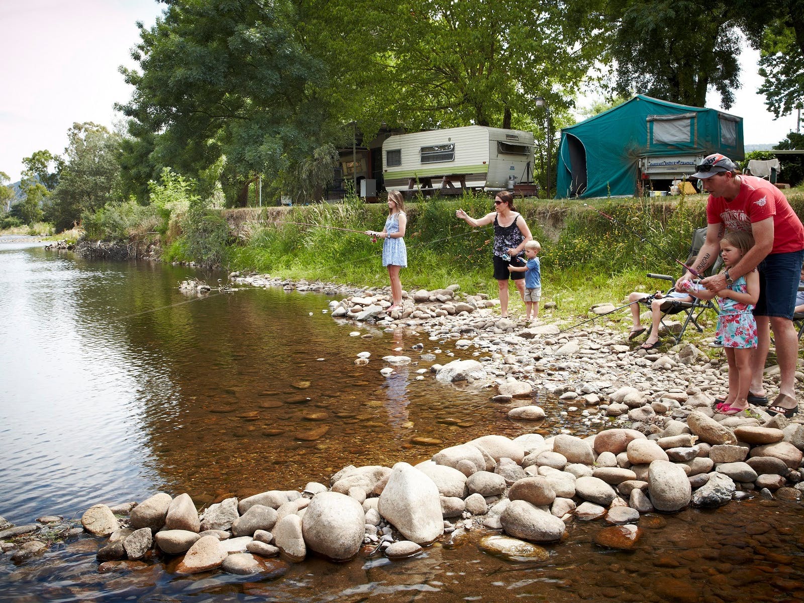 2 adults and 3 kids, fishing off the rocky bank by the river, caravans, tents, sunny day.