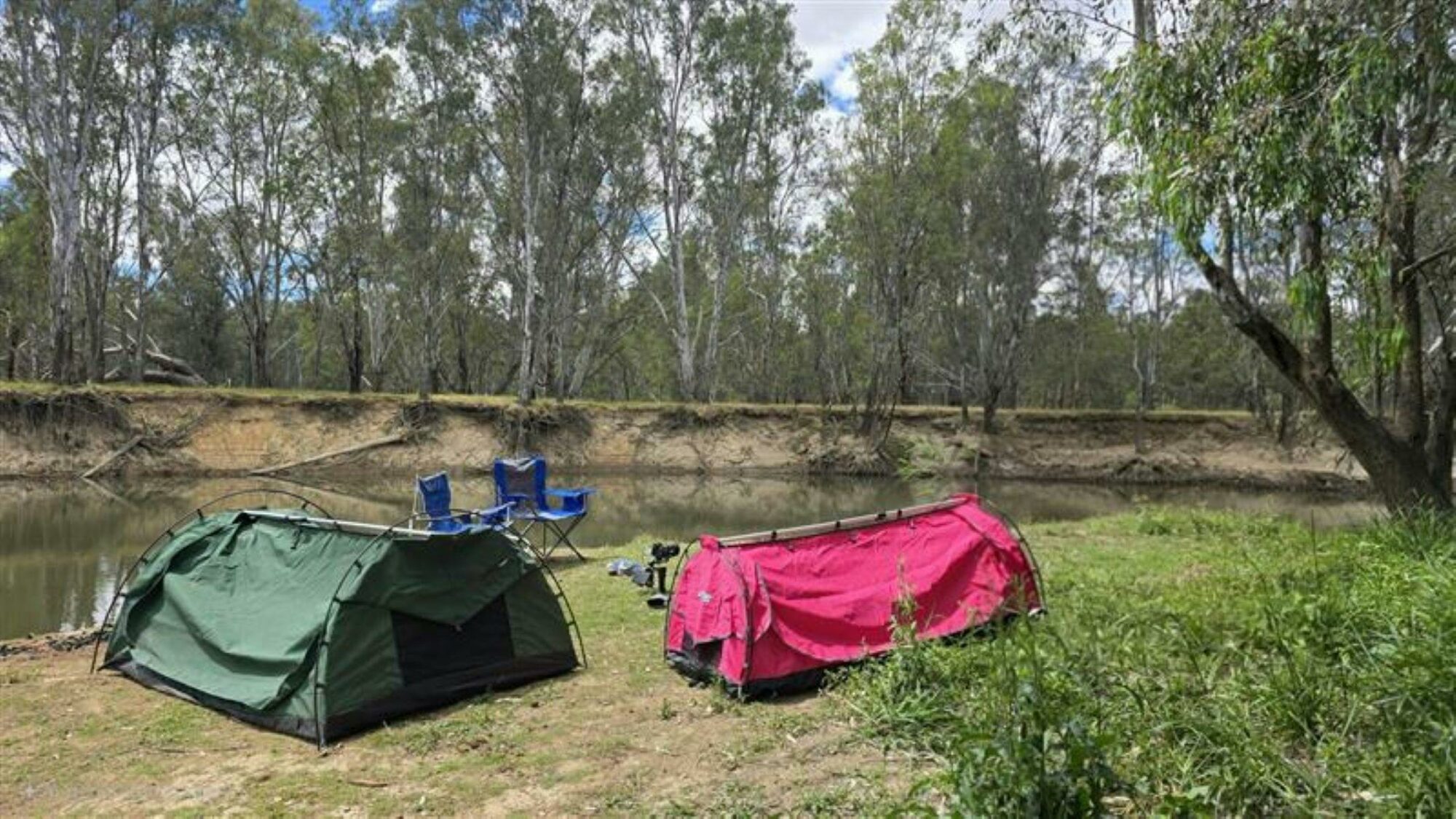 Green and pink swags set up on a grassy rivers edge with a river flowing behind the camp site.