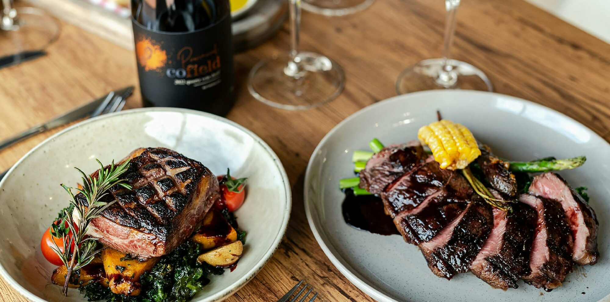 Two plated main courses featuring grilled beef, vegetables and red wine served on a wooden table.