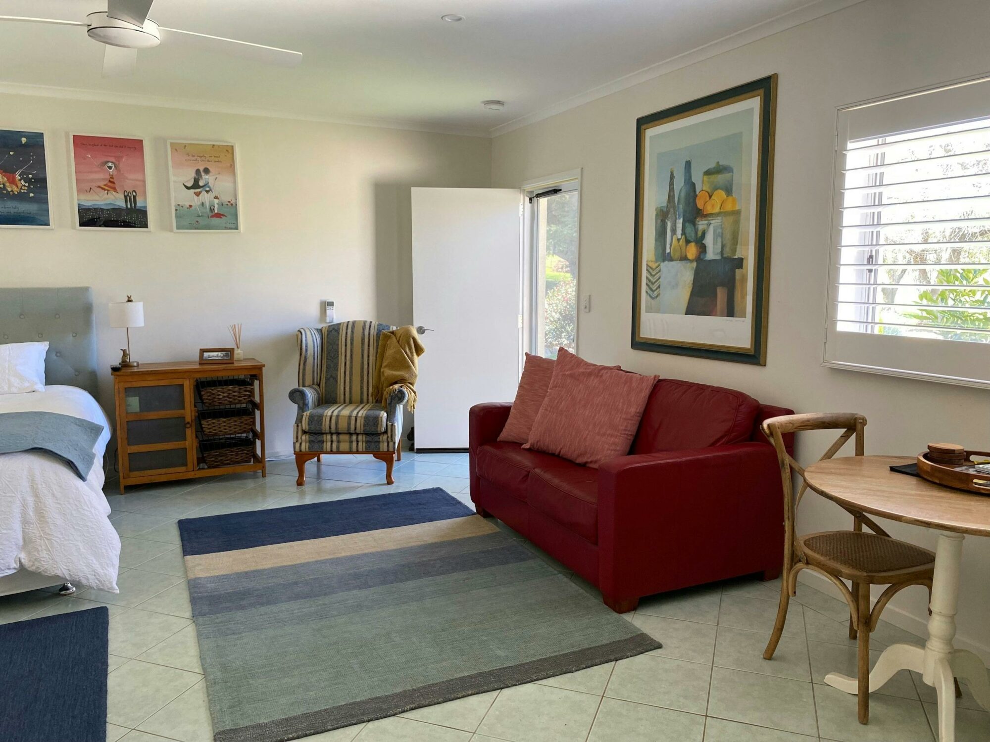 Red lounge with blue mat in front looking towards  bed. Arm chair near wall and bedside cupboard