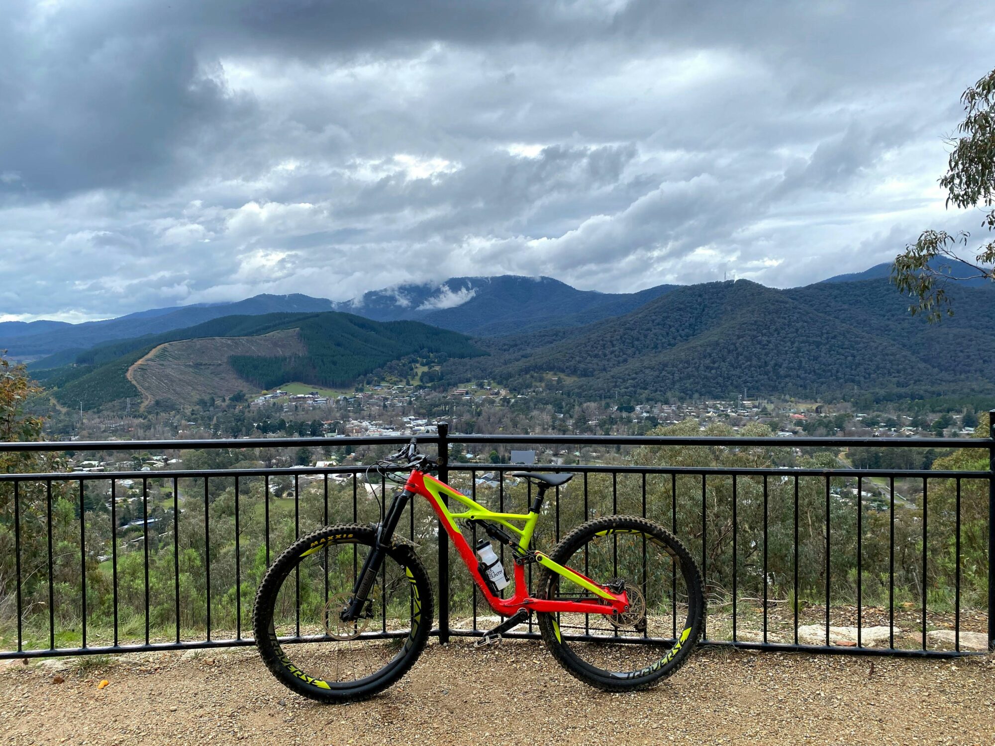 A red and yellow mountain bike leans against a fence with a view of mountains and towns behind.