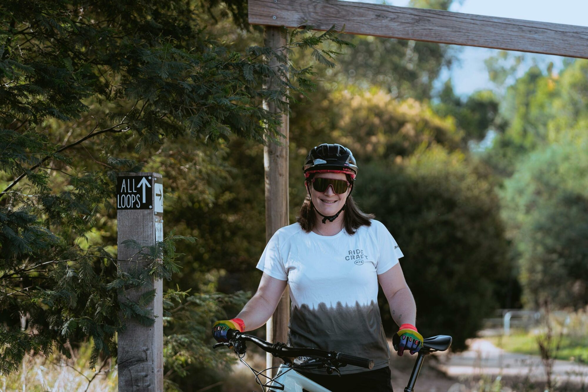 A rider in a white jersey stands with her bike in a forest.