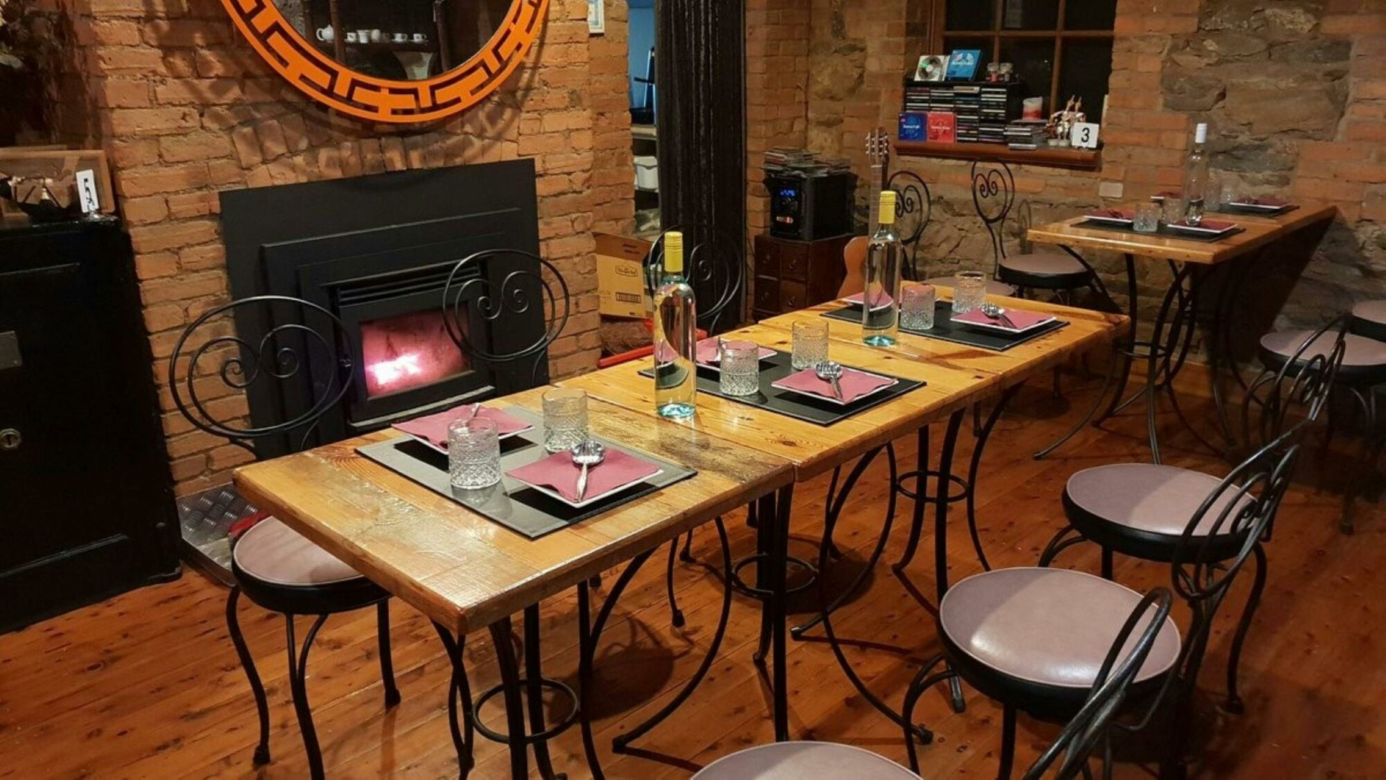 Timber table with 6 dining chairs, set with cutlery and glassware. A wood fire burns in background