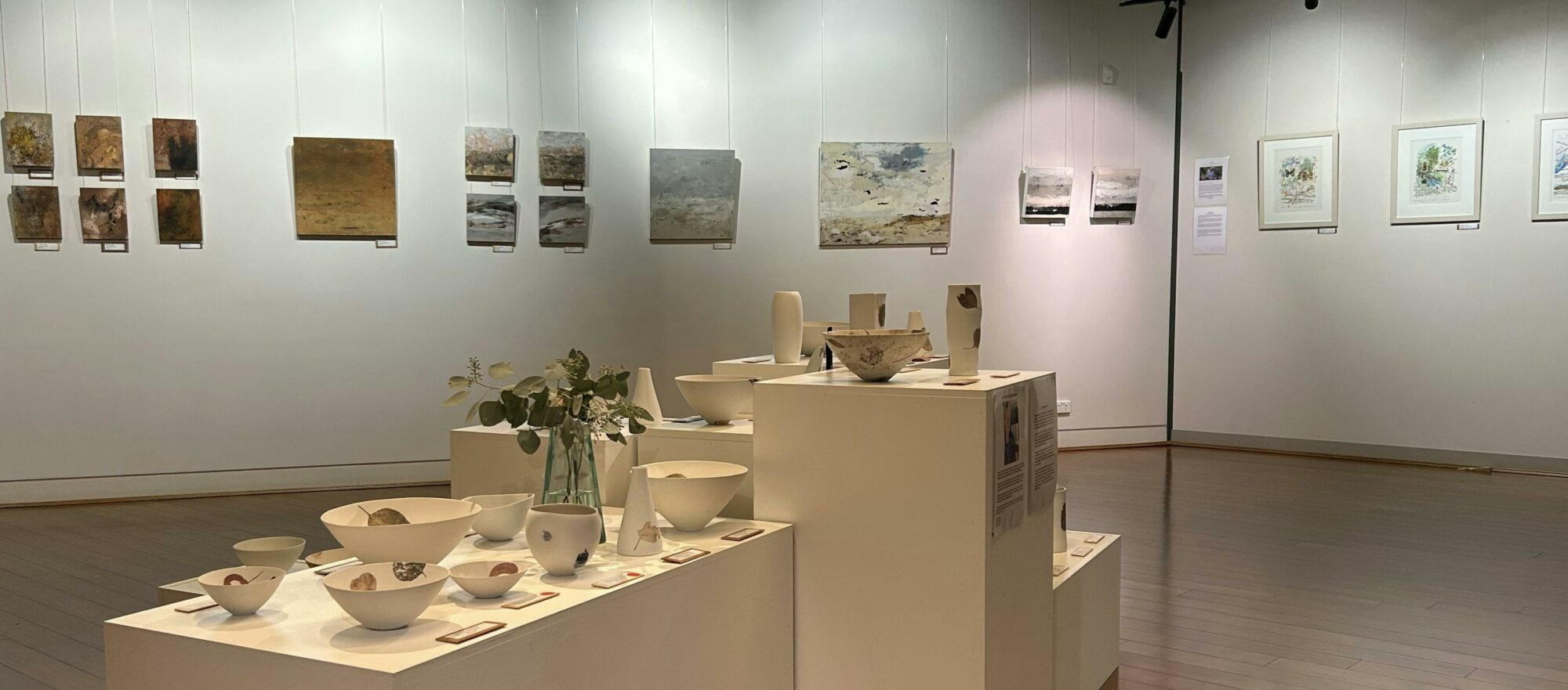 Mira gallery with paintings on white walls and pottery on white plinths in centre of room