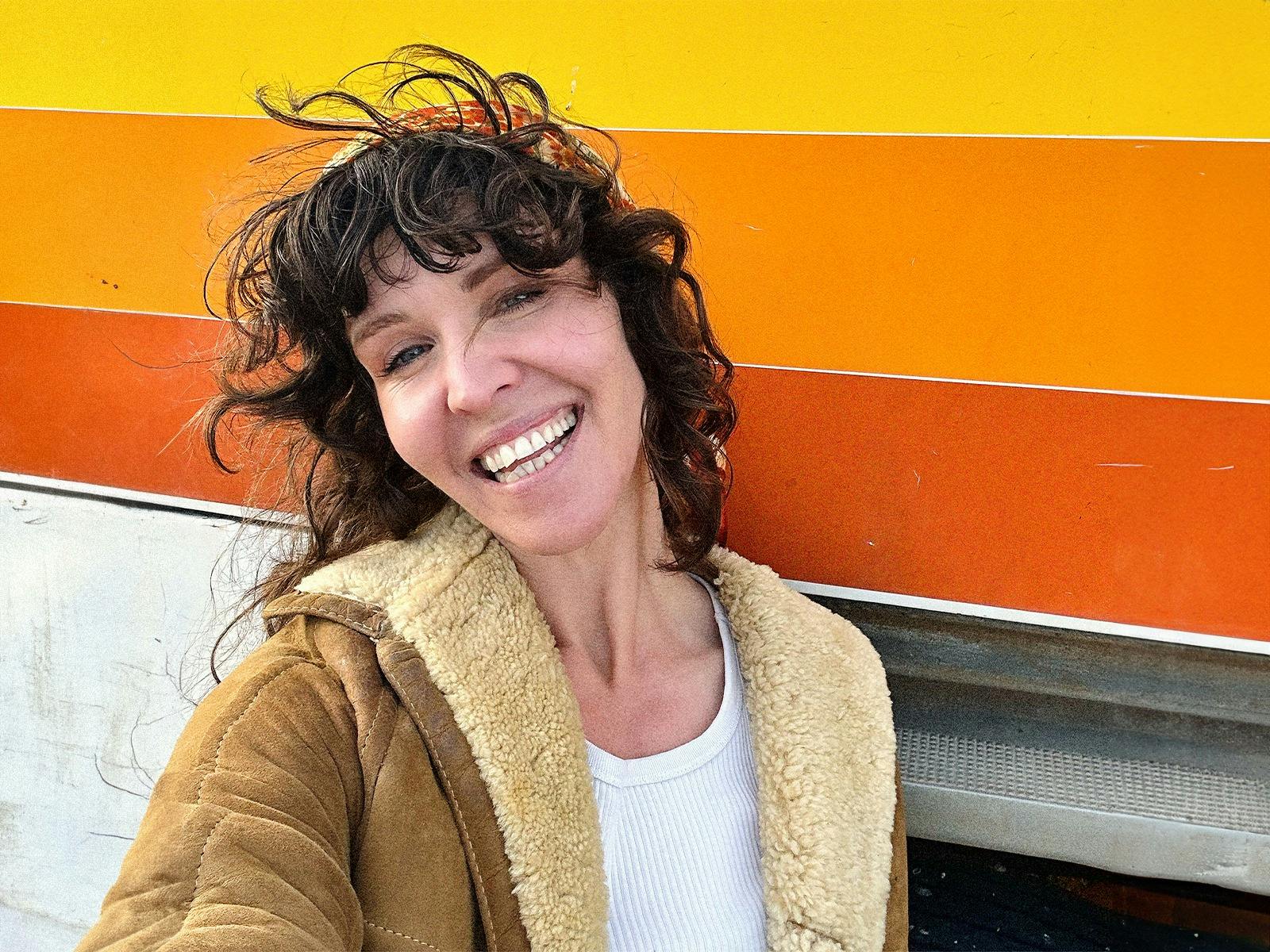 Smiling woman in sheepskin jacket with yellow, orange & ochre striped background.