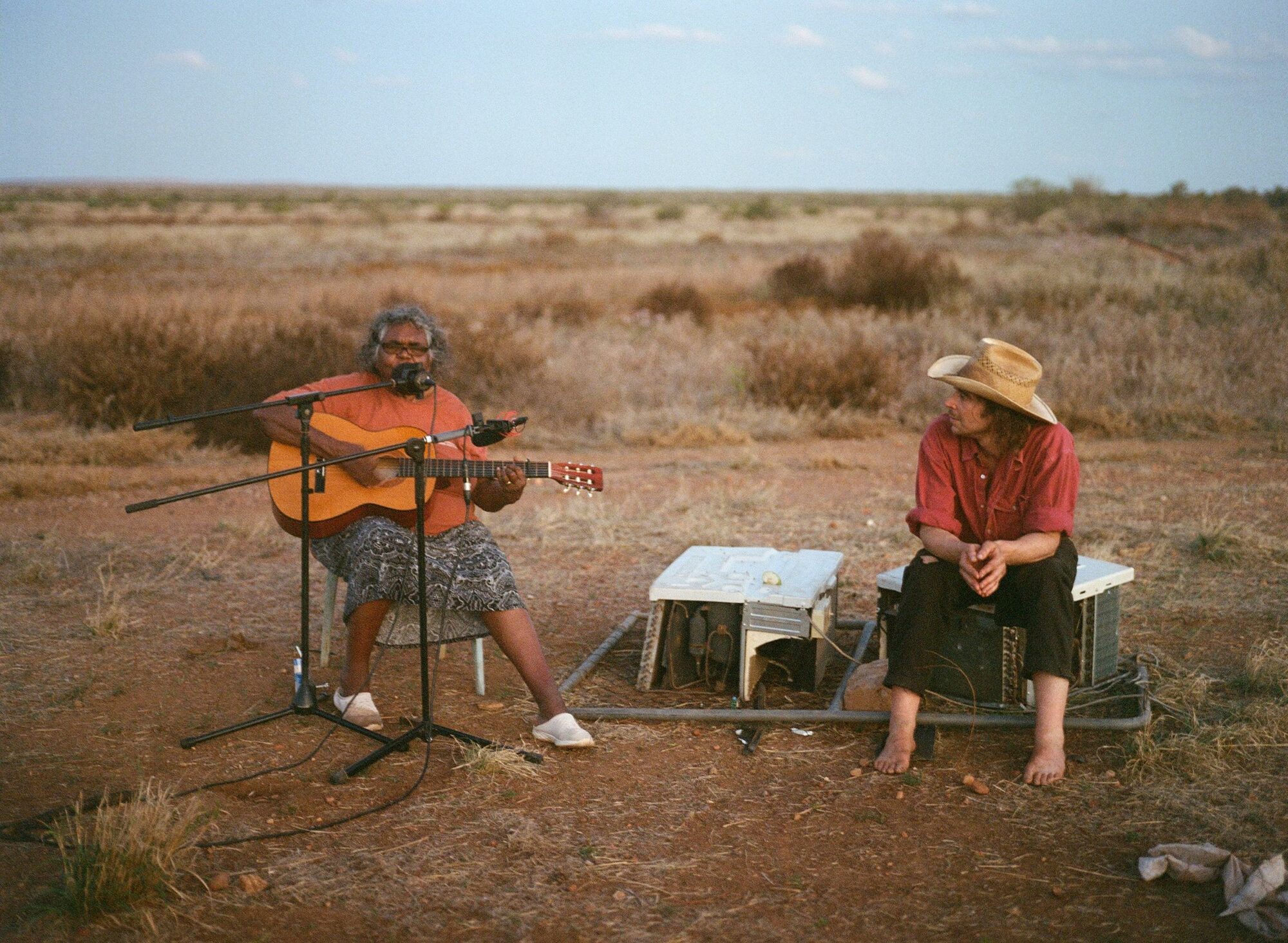 women sitting with guitar and man sitting next to her watching on. They are in a dessert landscape