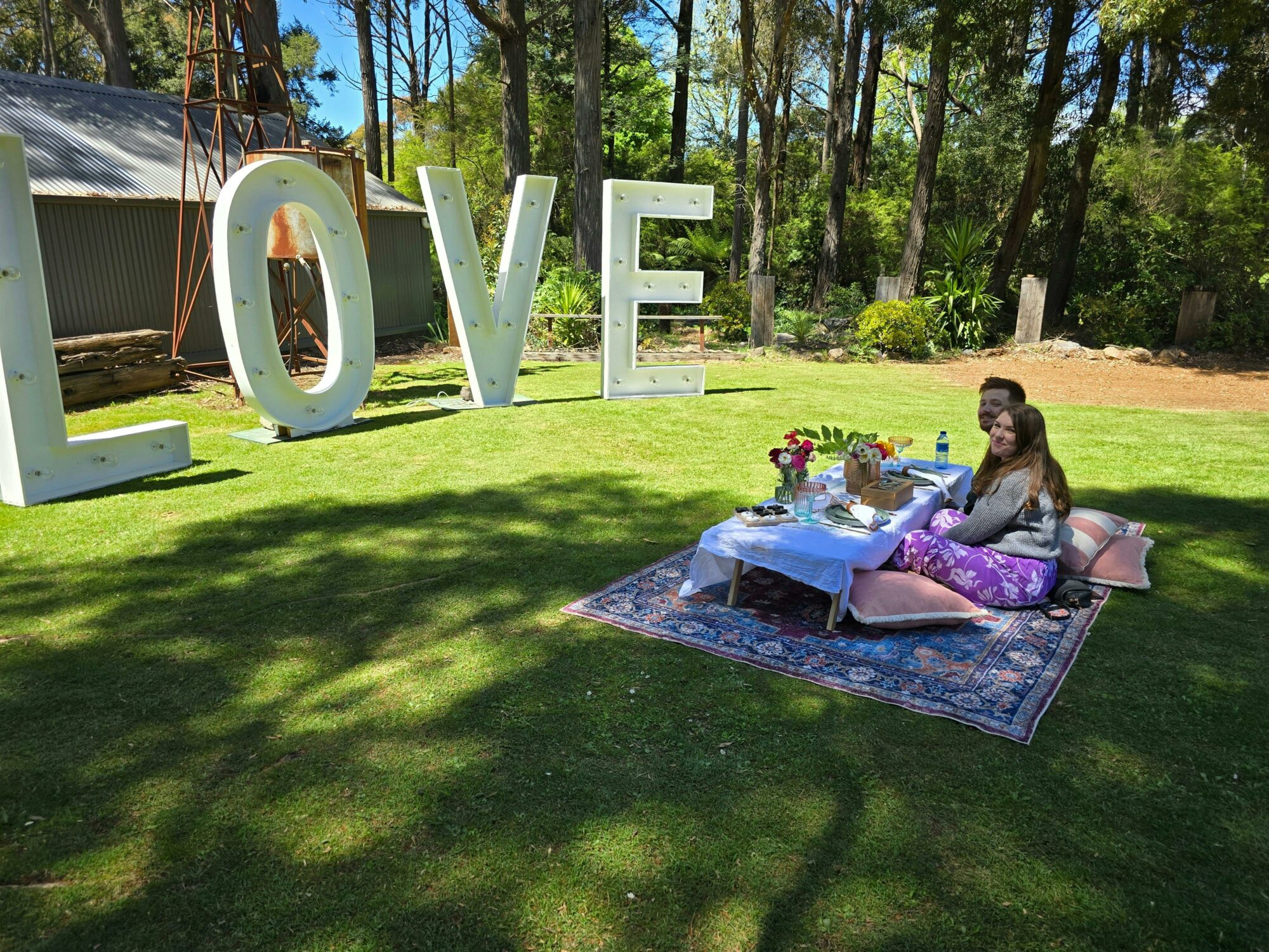 Photo of a man and woman sitting on a rug layed on a grassed area. A large love sign and picnic