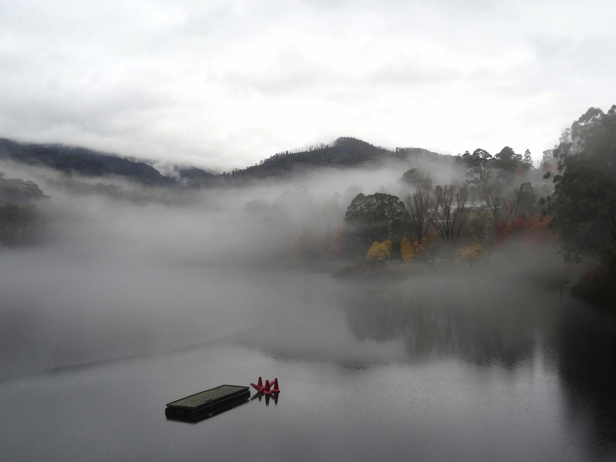 A  misty alpine lake with autumn trees, a floating platform, red cones, and faint buildings.