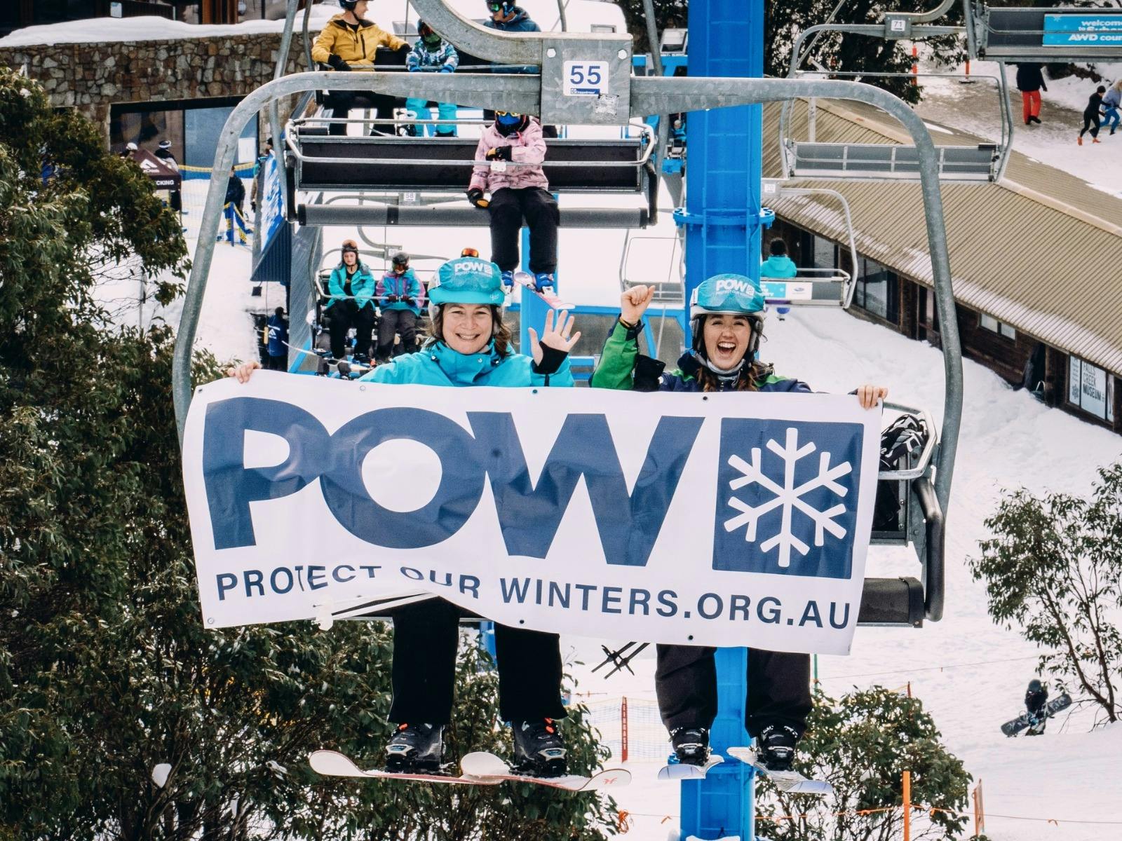 Two people sitting on a ski lift holding a sign