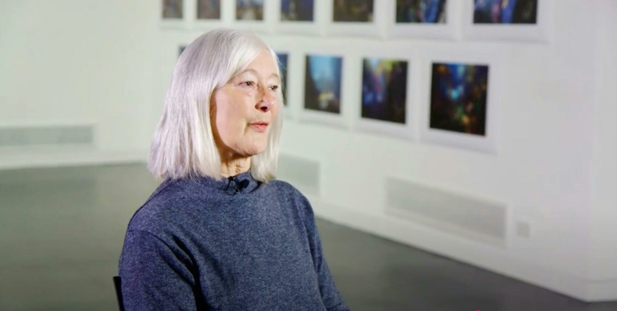 A woman with white hair speaks in an art gallery, with framed abstract artworks on the wall behind.