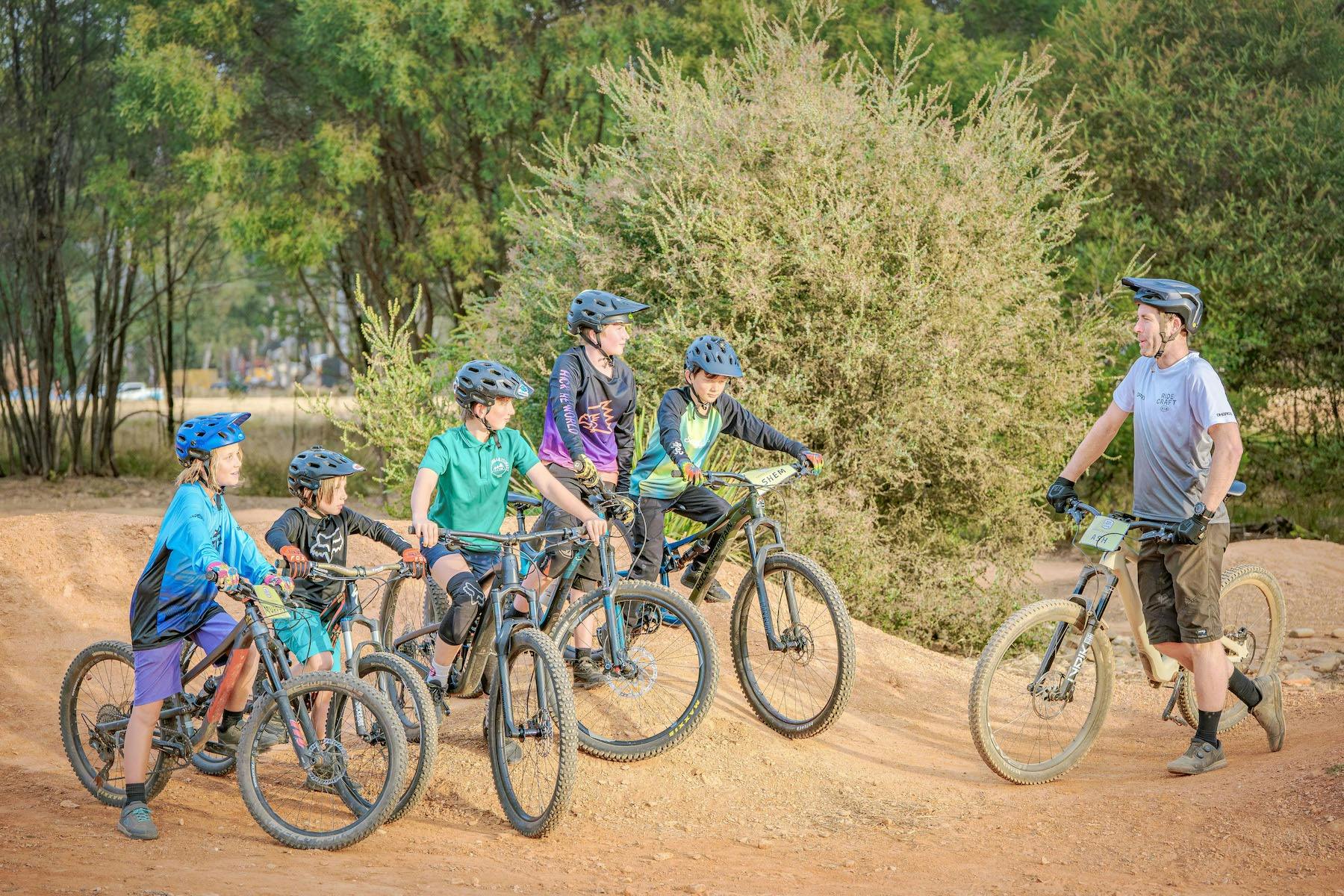 A coach with a bike stands facing a group of children with bikes in the forest.