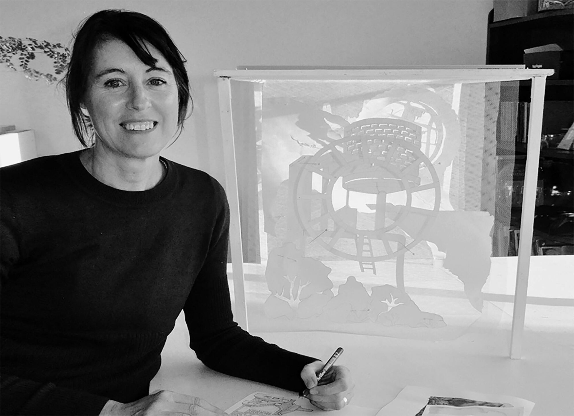 Artist Emma Ikin in her studio, smiling while sketching beside a delicate paper sculpture.