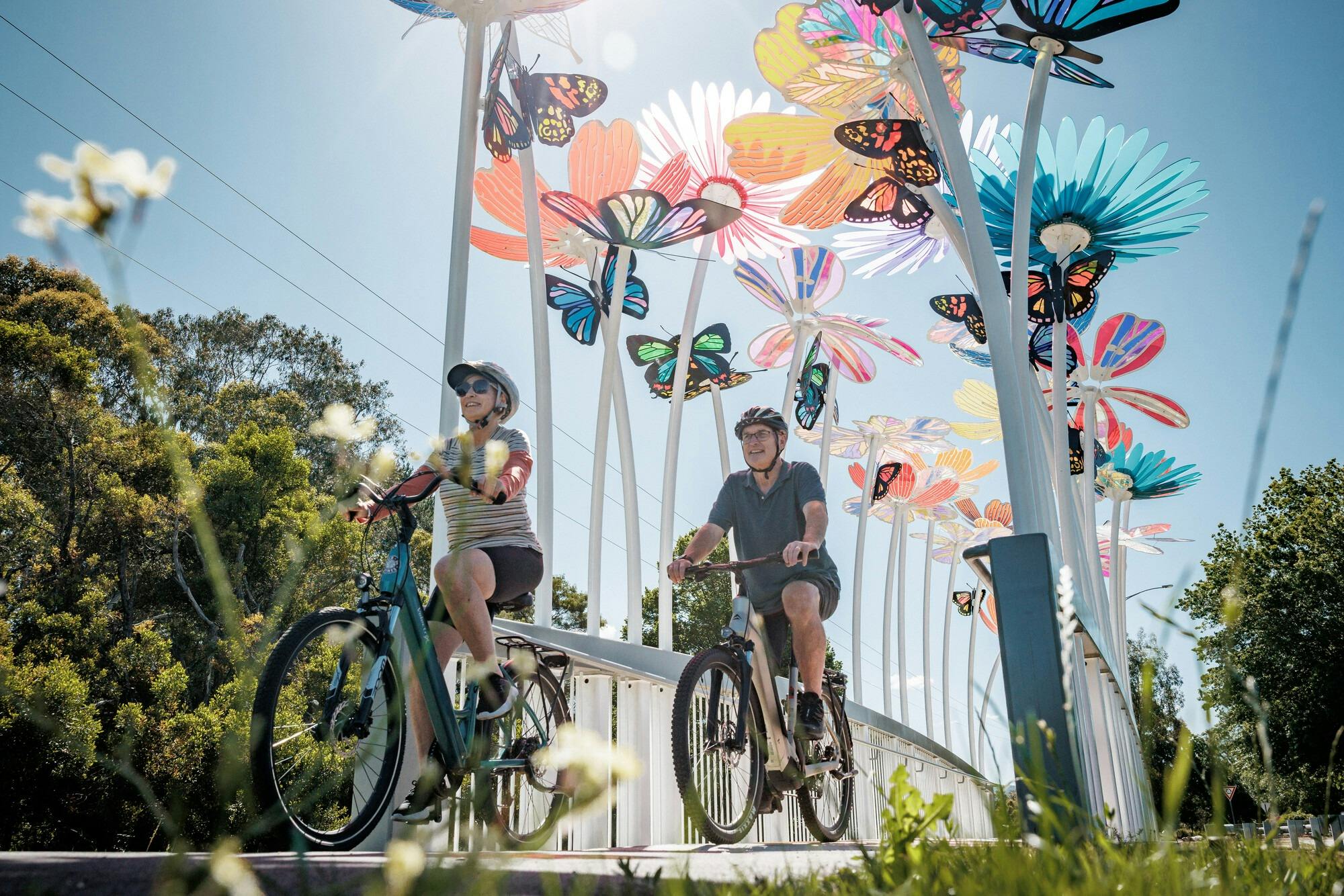 Two cyclists riding through a white, narrow bridge with tall colourful flower sculptures along it