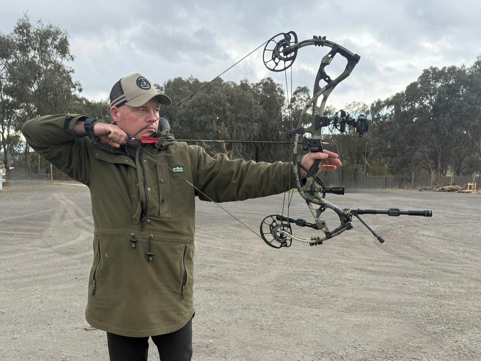 A man, Robert Deeble, wearing a Summit Outfitters Cap is holding a bow with arrow pulled back