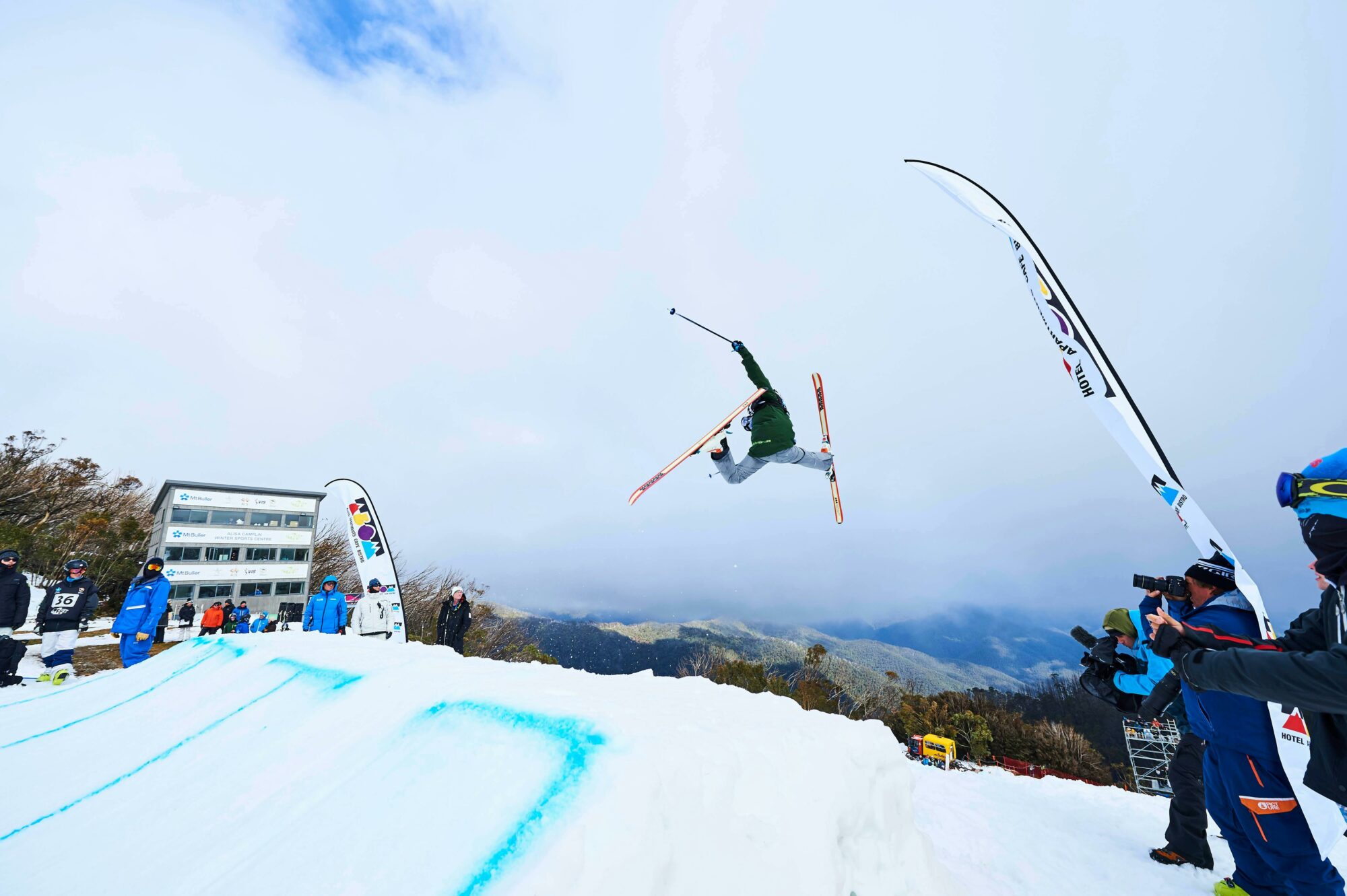 a skier in mid air with snow underneath them, and people on the ground holding cameras